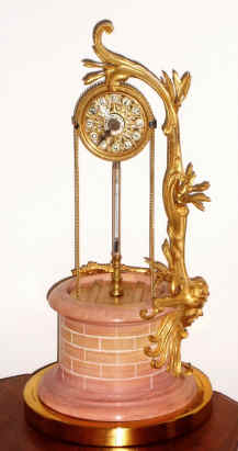 C.1890, French, Mysterious Timepiece, in the form of a wishing well, the dial indicated in the "Pulley" used to raise the water, and giving the impression that the chains turn the hands. Hands are actually driven by a shaft up the central support and through a pair of beveled gears. The leafy supports are all ormolu, the base of gilt brass. — Antique Clocks Price Guide