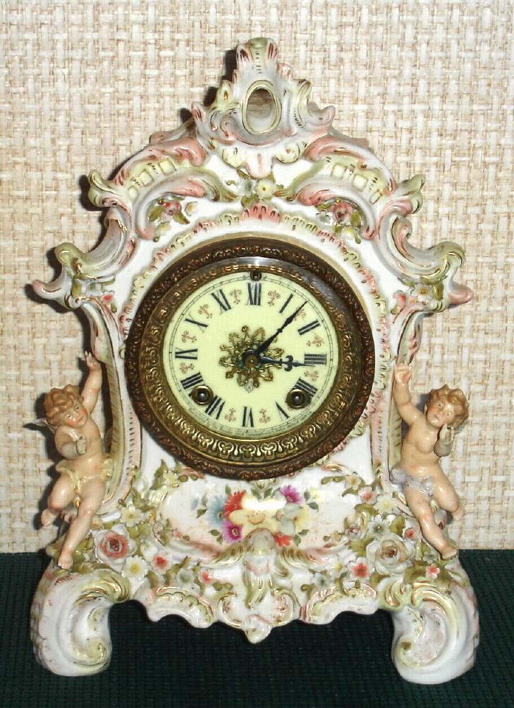 Ansonia Clock Co., unknown model, a European case in the French style with many 3-dimensional flowers with individual petals, the case with a winged cherub on each side with out-stretched arms, the porcelain case in light green tones with pink + gold highlights and multi-colored flowers. — Antique Clocks Price Guide