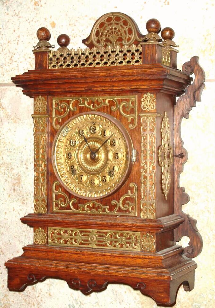 Ansonia Clock Co., New York, C.1894 "CABINET - F", antique oak cased mantle clock with brass trimmings. Has fancy embossed brass dial with raised Arabic numerals, This case with little brass fence or balustrade across the top , and with fancy brass bases to the ball finials. — Antique Clocks Price Guide
