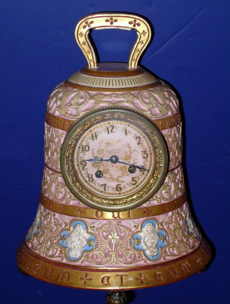 French, for Tiffany, Circa 1900, 8-day time and gong strike porcelain clock in the form of a church bell retailed by Tiffany and Co., NY. This is a porcelain version of the bronze "Pater Noster" clock that we have sold in the past {there is also a mini brass in tomorrow's session}. Has a fancy trademark of the porcelain company inside the case. The circumference has 8 angels in relief, each playing a different musical instrument. — Antique Clocks Price Guide