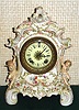 Ansonia Clock Co., unknown model, a European case in the French style with many 3-dimensional flowers with individual petals, the case with a winged cherub on each side with out-stretched arms, the porcelain case in light green tones with pink + gold highlights and multi-colored flowers.