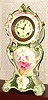 Ansonia Clock Co., Royal Bonn, "No. 506", slender porcelain clock in the French style, with one piece porcelain dial, creamy colored with red accents between the numerals; the white case with misty green edges and dark green accents + gold highlights, and a large cameo with floral spray below the dial. Sides of case decorated with violet vines.
