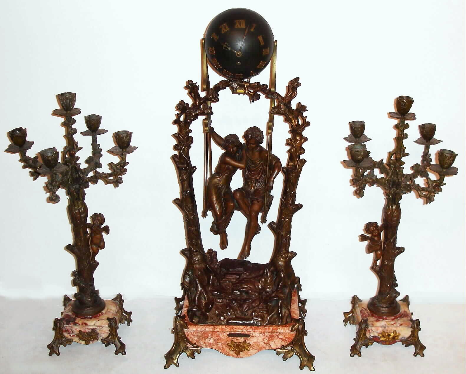 French, very large swing clock, 3 piece set with matching 5 light candle stands. A romantic couple are seated in a swing, appearing to suspend by rope from entwined branches joined at the top, and with a brass title plaque: "Idylle Printaniere, par Geo Maxim", the couple swinging from side to side without apparent impulse, C.1890 — Antique Clocks Price Guide