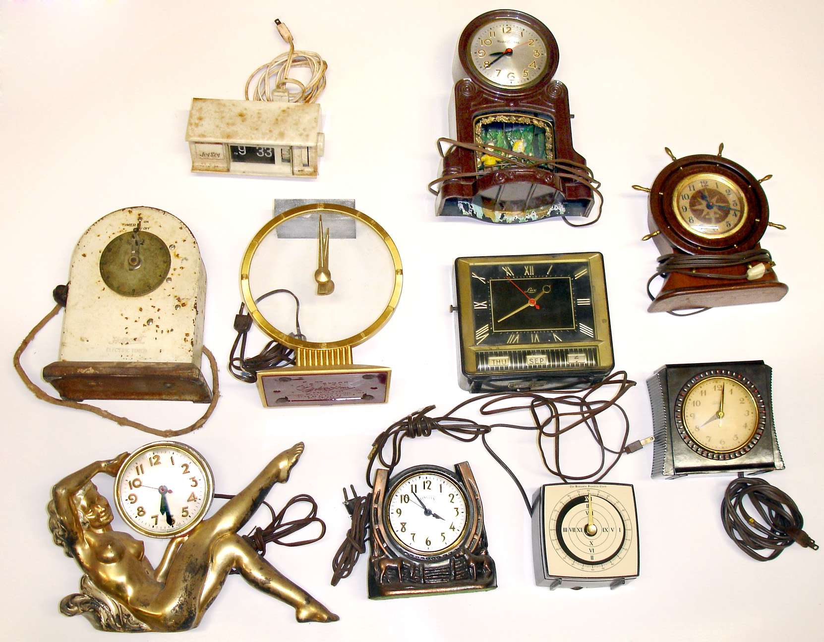 Ten plug-in electric clocks. Includes Golden Hour, Seth Thomas ship-wheel, Lux calendar, Benjamin Franklin and Hotpoint timers, Mastercrafters swinging dolls, reclining nude cast-metal, Jet-Set digital, Telechron appliance switch, and Lincoln horseshoe. — Antique Clocks Price Guide