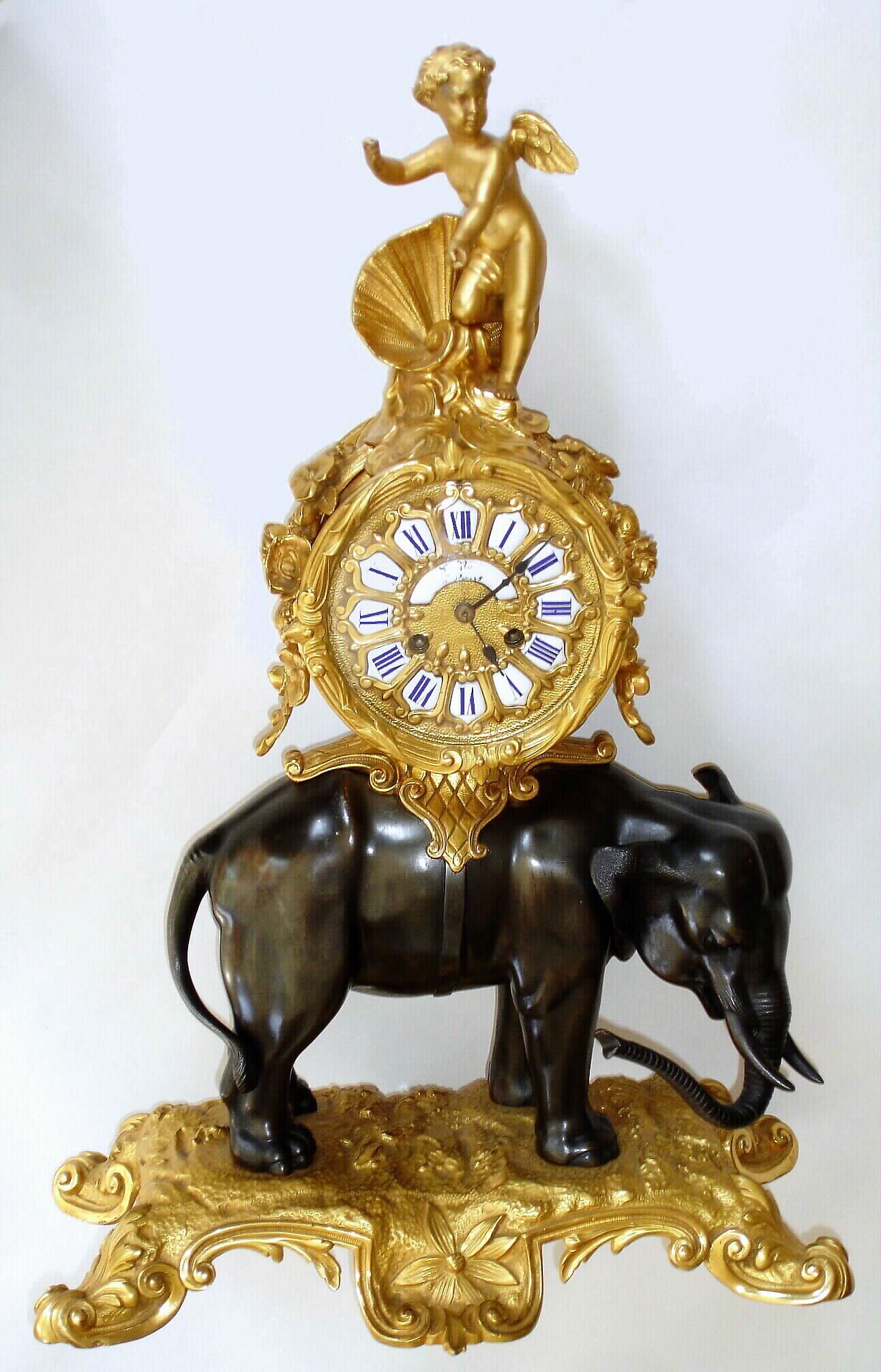 French, L'Elephant, in the style of a century earlier, bronze and ormolu, the clock mounted above the elephant, surmounted by a winged cherub. The pendulum extends from the movement down through a hole in the top of the elephant (original) and saddle , thus invisible from the outside. The dial with royal blue numerals on fired porcelain cartouches. Original name on dial not legible, however Paris can be read. Case components bear 3 letters L.L.A., c1870 — Antique Clocks Price Guide