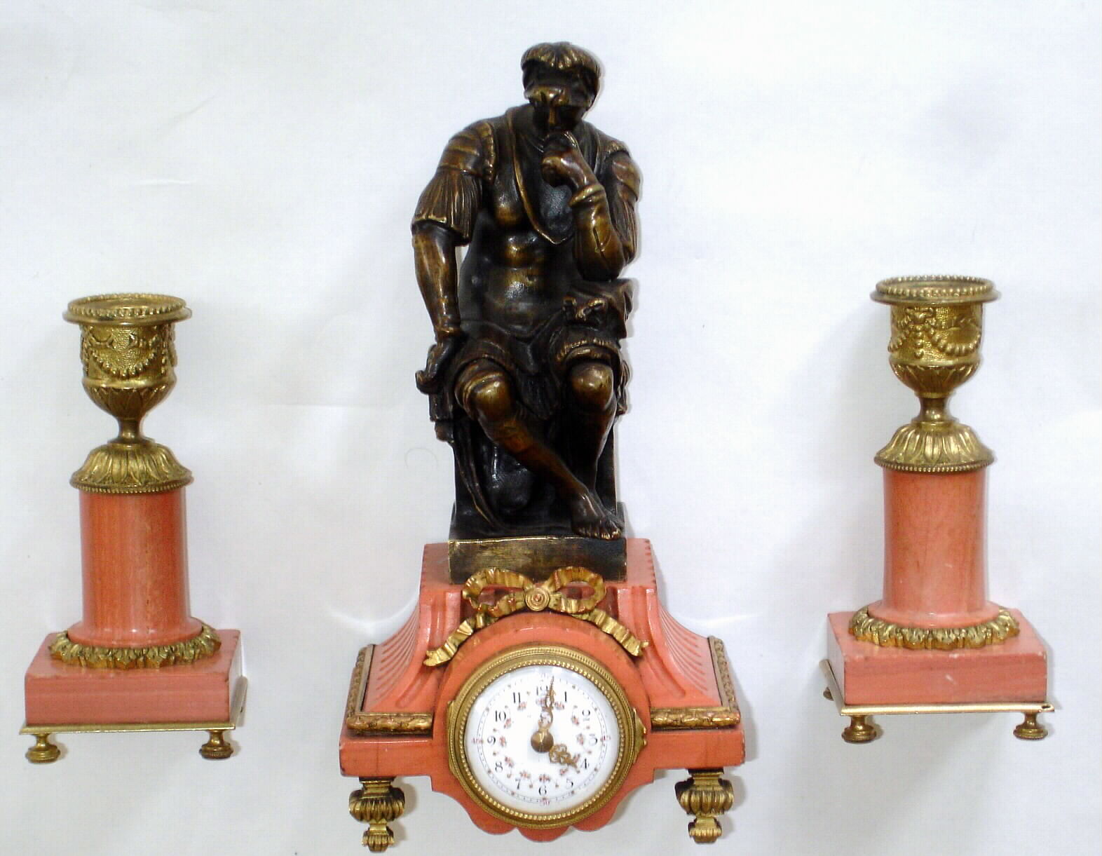French, miniature 3-piece garniture desk set, salmon colored marble and bronze figure with ormolu trim, and with a tiny round French movement and fancy porcelain dial, and tiny pierced and chased gilt hands. Note seated warrior in tunic is well detailed and he wears a helmet of tooled leather over the top of a bear's head, c1880 — Antique Clocks Price Guide