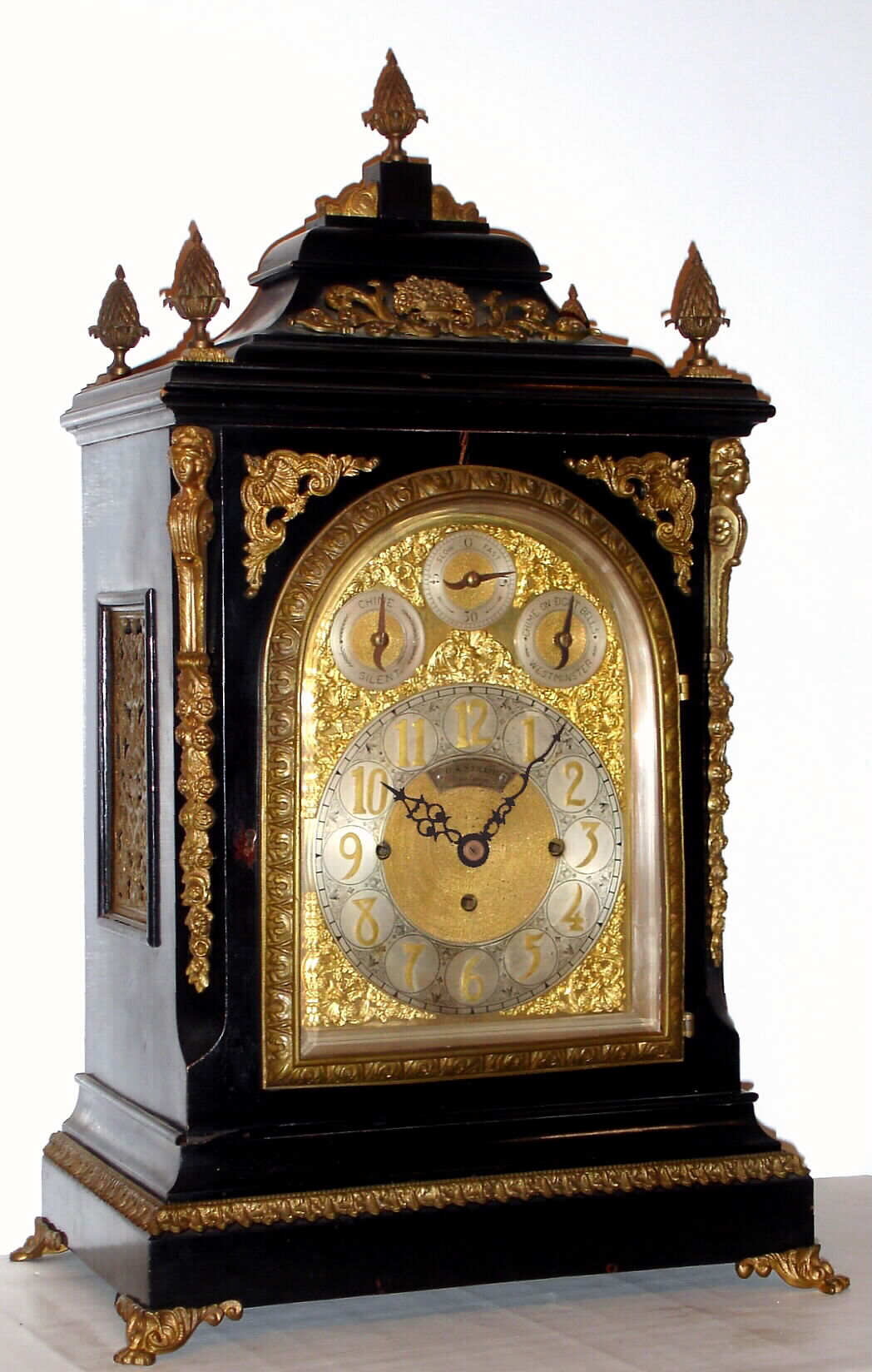 English, triple fusee, quarter chiming bracket clock, in a classic Georgian style large ebonized case with brass caryatids, side frets and pineapple finials, the whole resting on cast "toed" feet, chiming your choice of Whittington on 8 bells or Westminster on 4 large flat wire gongs, and then sounding the hours on a still larger steel flat wire gong, the clock retailed by O. A. Strapp, Cambridge, and with an engraved cartouche below 12 bearing that information, and the arched dial with small subsidiary dials labeled Chime /Silent, F/S, and Chime on 8 Bells or Westminster, C.1875 — Antique Clocks Price Guide