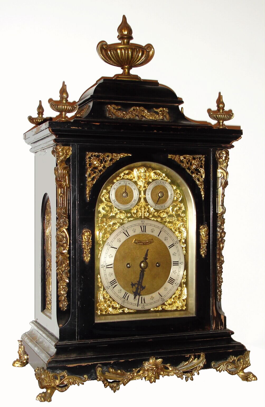 German {in the English style} large fusee bracket clock, retailed at 29 Church St, Liverpool by Frisch Schierwater and Co (on dial), an ebonized case with cast brass "jester" caryatids to the corners, fancy cast brass side frets, all surmounted by a giant urn and flame finial, with smaller finials to the corners, the whole resting on "toed" feet. Note this clock chimes Westminster quarters on 4 tuned flat wire gongs and strikes the hours on a much larger 5th deep toned gong. Subsidiary dials on the arched dial are for Fast / Slow and Chime / Silent. The dial with cast spandrels of winged cherubs on both the dial corners and at the apex of the arch, and gilt foliate castings filling in other areas. Note this is a two [2] fusee clock, with reliable quarter chime and strike system designed by Winterhalder and Hofmeier....see horizontal pins on rear of tune drum to perform strike. C.1890 — Antique Clocks Price Guide