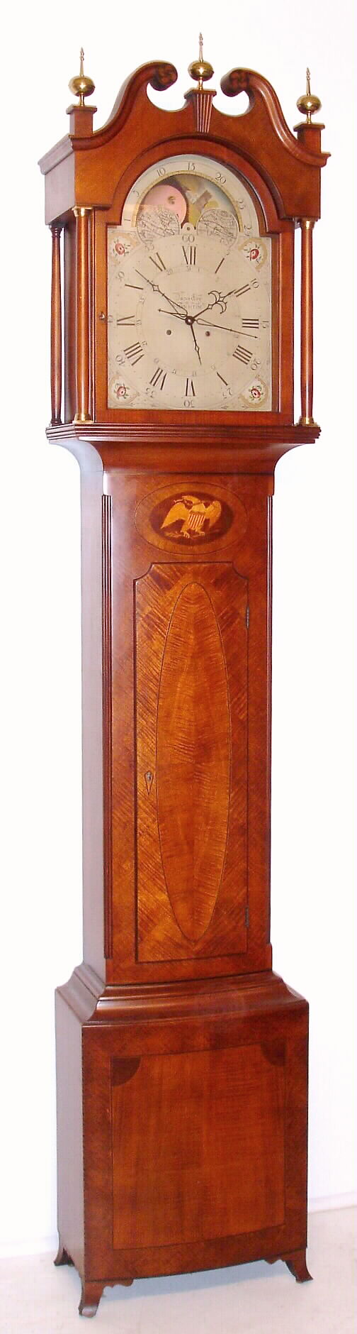 Colonial Clock Co., Zeeland, MI, reproduction of a Pennsylvania tall clock by Jacob Eby of Manheim, PA, time and bell strike West German mov't, in a bowed front tiger maple case and painted metal dial with moon phase, calendar and center seconds hand. No. 120 of a limited edition of 300, This model was the most popular of the 6 styles produced; they made each of the models that stood in the front hall of the Henry Ford Museum at the time. C. 1975 — Antique Clocks Price Guide