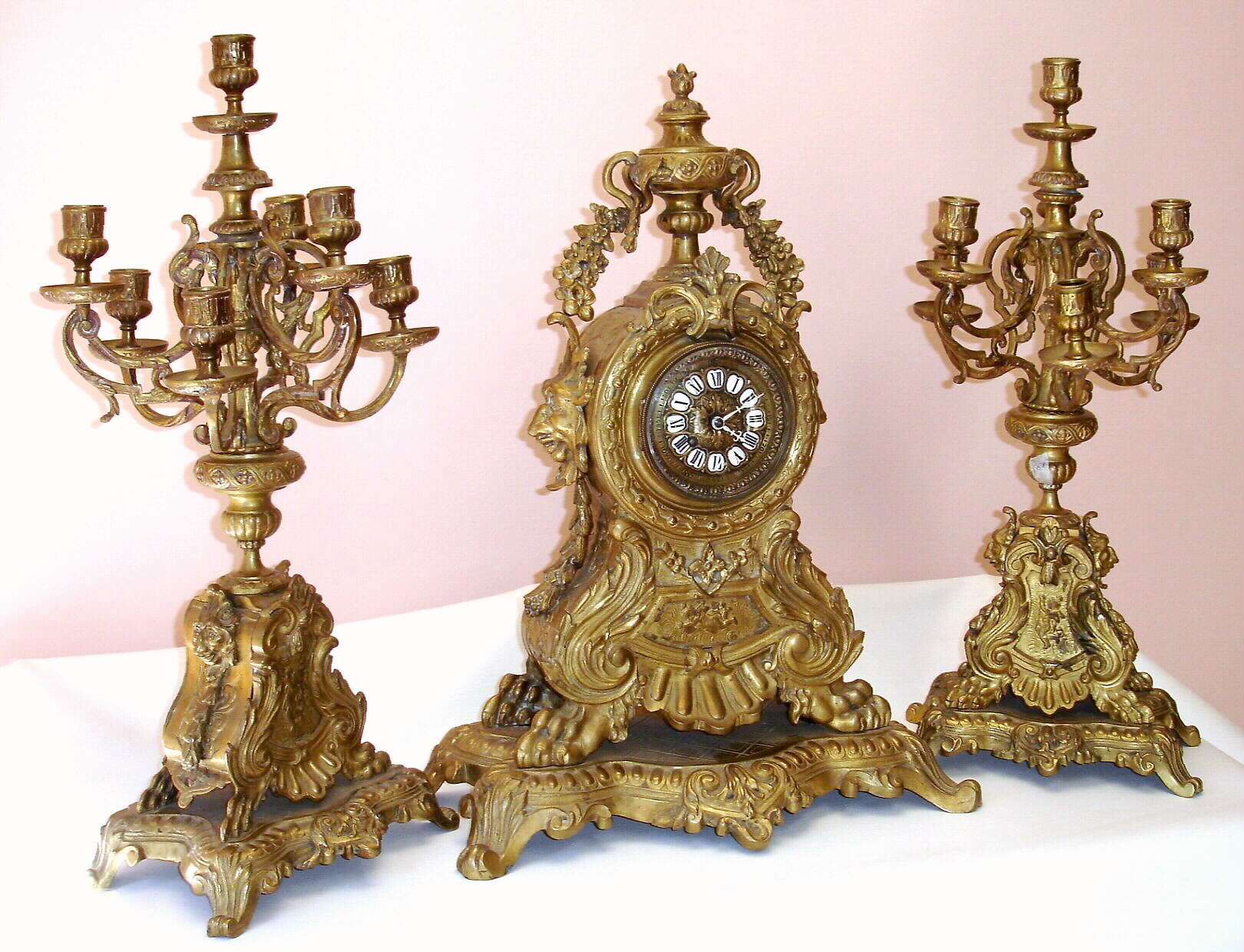 French, large cast brass three piece garniture set with matching candelabras. The cast dial has 12 individual cartouche numerals, and pierced steel hands, now white. Case bears garlands and shell motif and large lions heads flank the clock and smaller lions heads flank the candle stands, c1890 — Antique Clocks Price Guide