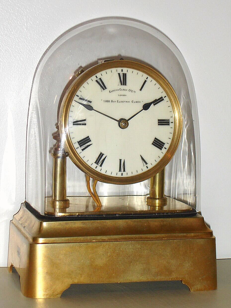 Eureka Clock Co, London, "1000 Day Electric Clock, electro mechanical mantel clock, battery powered, time only with large cut bimetallic balance and blued steel balance spring, Roman numeral white enamel dial with heavy gilt bezel, blued steel hands, movement with spotted back plate, supported on gilt columns, with finial style nuts securing the movement, all on a gilt brass base, and covered with a glass dome, s#14614 c1910 — Antique Clocks Price Guide