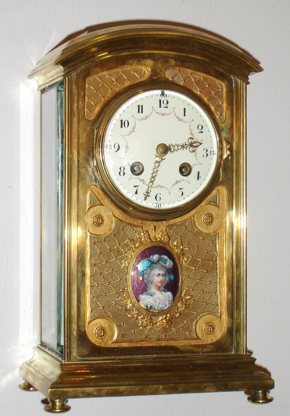 French crystal regulator in fancy polished brass case, spring powered time and strike movement, heavy beveled glasses on sides, back and top, the front with latticework panels backed with brass, decorated in a moire pattern, with an enamel portrait of a lady in formal attire below dial, on a transparent violet enamel background, showing a moire patterned ground beneath, molded bezel with beveled convex glass, Arabic numeral white enamel dial with flowered swags draped at each numeral, excellent pierced and engraved brass hour and minute hands, c1910 — Antique Clocks Price Guide