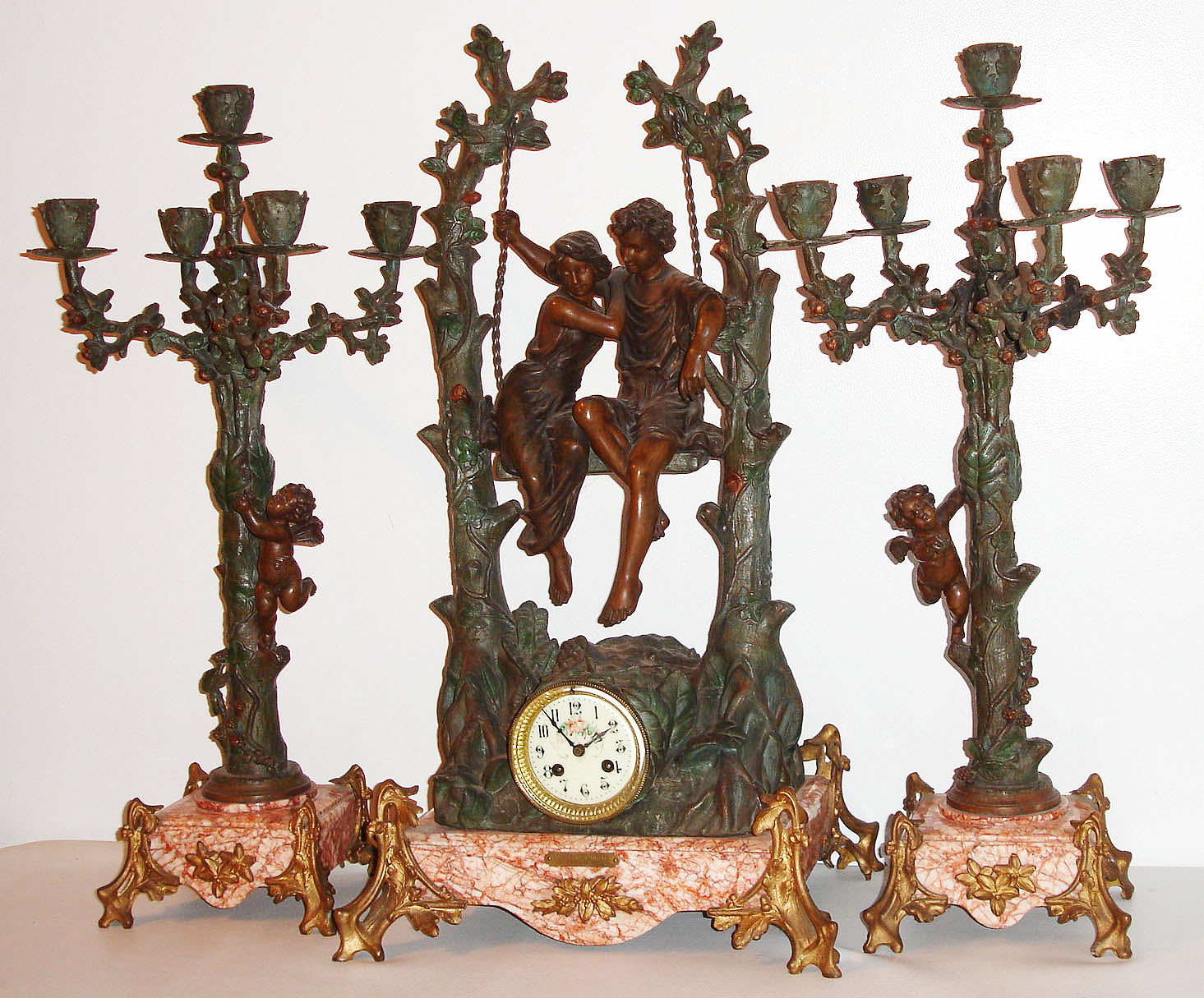 French figural garniture with candelabra, with 8 day pendule a Paris time and strike movement, recoil escapement and count wheel striking on a bell, white enamel Arabic numeral dial with floral decoration, blued steel hands, gilt brass bezel with beveled glass, the large case of patinated spelter, with a young couple swinging between two pruned tree trunks, the trunks supporting vines with leaves and ferns below, the rustic style feet with oak leaves and acorns, on a marble base with fine red orange veining, and set with a small plaque engraved "Idylle Printaniere Par Geo. Maxime", the right side of the clock case also signed Geo. Maxime, the candelabra also with pruned trunks supporting vines, each with a single Putti, ending in spreading branches with leaves and budding flowers, each supporting one bud form candle cup, and supported by marble bases on 4 rustic feet, with key c1880 — Antique Clocks Price Guide