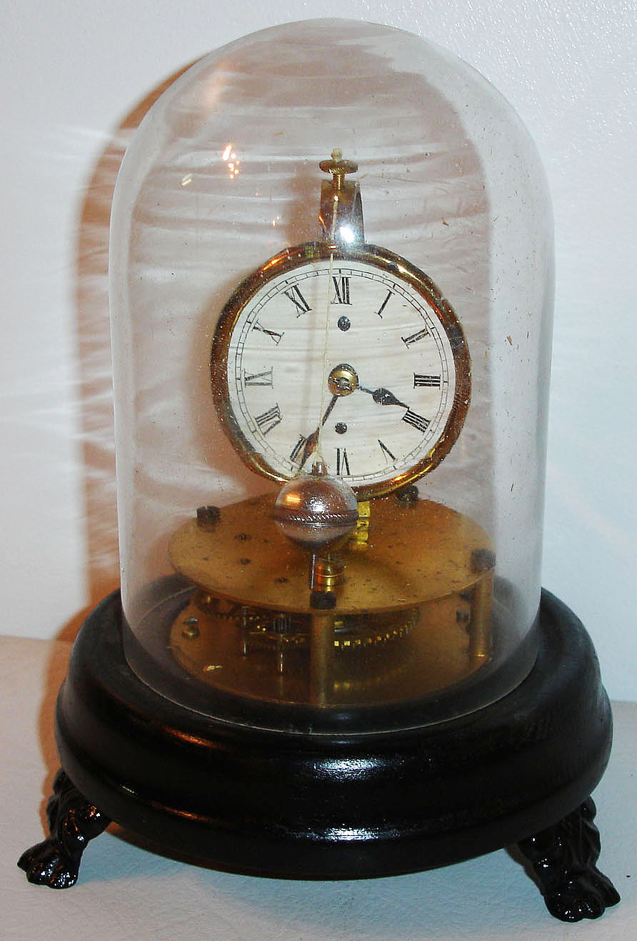 E. N. Welch Mfg. Co., Briggs rotary pendulum novelty clock (30 hour), black painted round wooden base on cast iron paw feet, supporting a horizontal round plate movement, with vertically mounted Roman numeral dial printed on glossy card stock, blued steel hands, and rotating pendulum hung in front of the dial, under a glass dome c1875 — Antique Clocks Price Guide