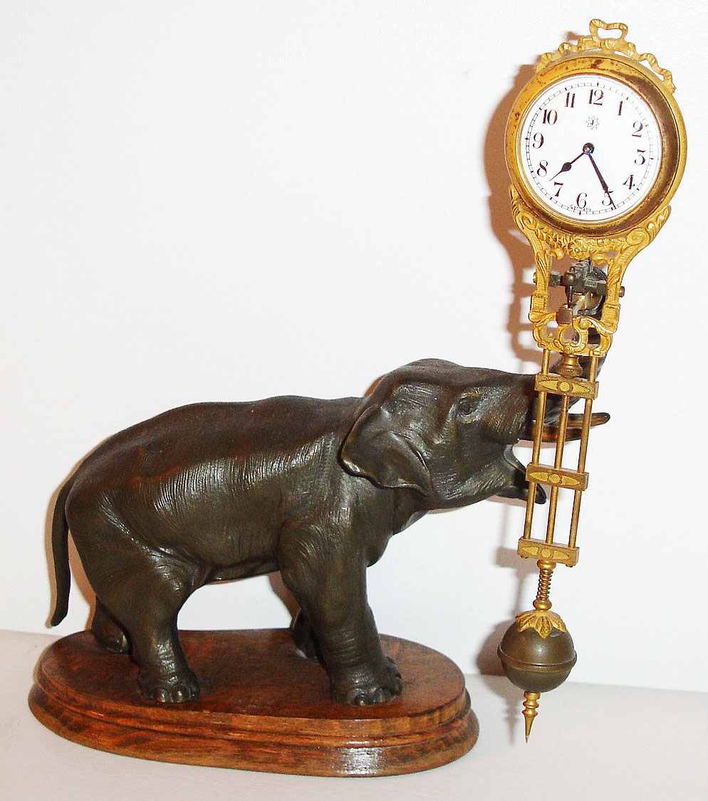 Junghans, Germany, "Elephant" model swinger, 8 day novelty timepiece, the patinated spelter elephant mounted to a molded beech wood base, his trunk supporting the gilt and patinated clock case/ pendulum, white enamel Arabic numeral dial, blued steel hands, ca.1920 — Antique Clocks Price Guide