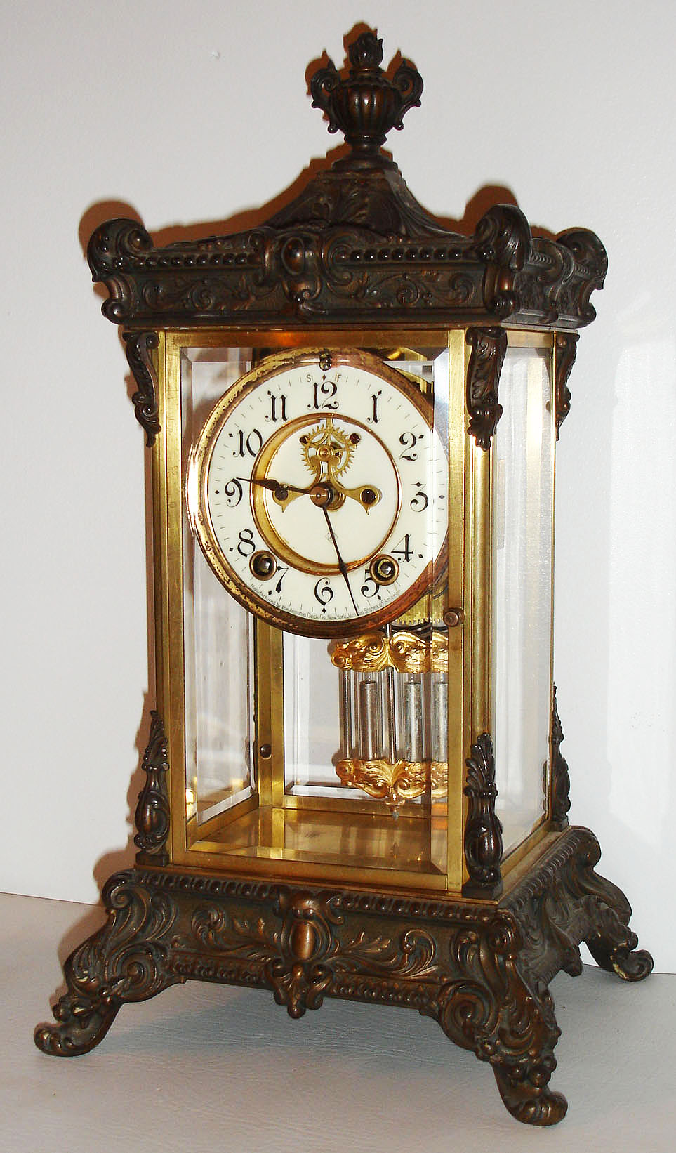 Ansonia Clock Co., Brooklyn, N.Y., "Elysian" model crystal regulator, ornate cast top and bottom with gilt finish, beveled glasses, white enamel Arabic numeral dial, 8 day, time and strike, movement with round plates, Brocot open escapement, gilt simulated mercury pendulum c1915 — Antique Clocks Price Guide