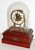 Eureka Clock Co., Ltd, London, England, the "1000 day" battery powered movement with large bimetallic balance and spotted brass plates, supported by two turned baluster form columns, with white enamel Roman numeral dial, blued steel spade hands, brass bezel with convex beveled glass, all covered with a glass dome, and mounted to a beautifully made, polished mahogany base with battery compartment, the front with a brass plaque engraved "Eureka 1000 Day Motion" c1910
