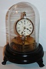 E. N. Welch Mfg. Co., Briggs rotary pendulum novelty clock (30 hour), black painted round wooden base on cast iron paw feet, supporting a horizontal round plate movement, with vertically mounted Roman numeral dial printed on glossy card stock, blued steel hands, and rotating pendulum hung in front of the dial, under a glass dome c1875