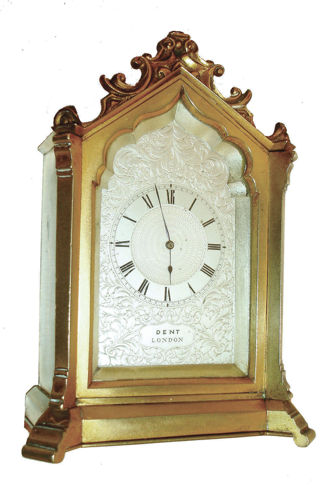 Dent, London, 8 day time only, desk clock in the Gothic style, the gilt case with stepped base and molded feet, gable with applied scroll ornament, modified lancet arch front with conforming, silvered dial mask framing a foliate engraved silvered Roman numeral dial, the center with engine turning, and signed "Dent London" below the chapter ring, blued steel spade and poker hands, small French style carriage timepiece movement with lever platform, case back with apertures for regulation, setting and winding, with shutter for exclusion of dust, and marked "3038", stamped crown over a lozenge shaped reserve marked "E.W.S.", an hourglass, and banner with "Machine Made", c1870 — Antique Clocks Price Guide