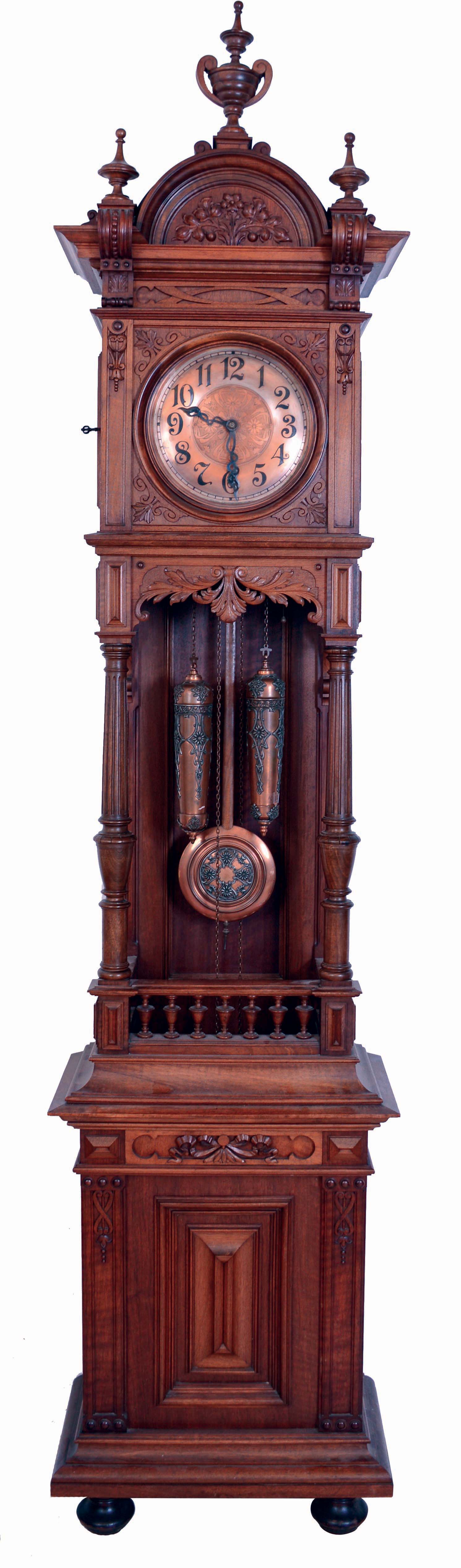 Furtwangler, L. & Sohne, Furtwangen, Germany, open well tall clock, 8 days, time and strike, on gong, brass weight driven movement in a carved walnut case on bun feet with large turned finials and copper finished brass dial. The dial, pendulum bob and weights are matching filigreed copper tone, serial #13235 — Antique Clocks Price Guide