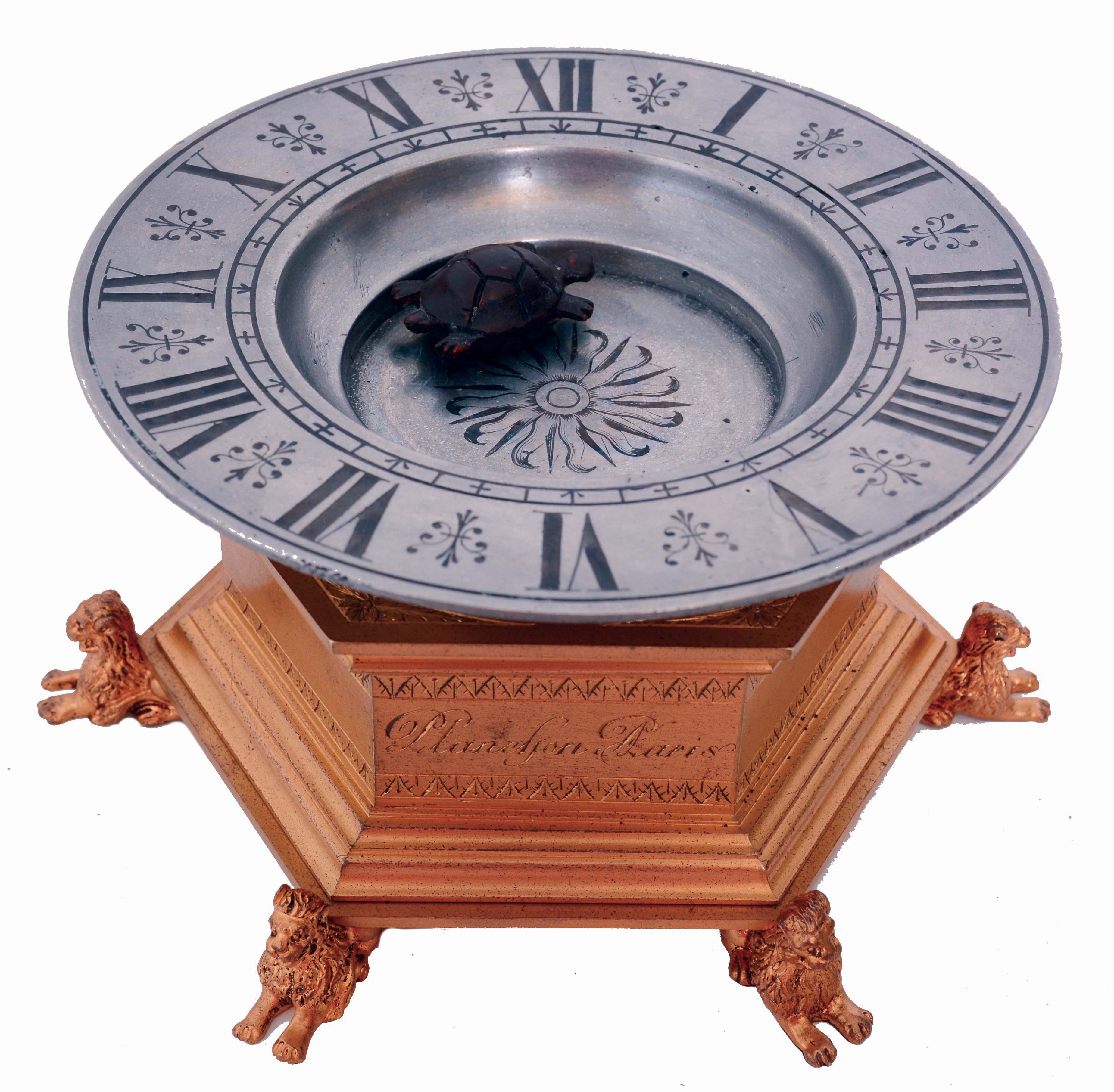 Planchon, a Paris, turtle mystery clock of small size, the molded hexagonal, gilt bronze base with decorative engraving resting on recumbent lion form feet, supporting a pewter basin with Roman numeral chapter engraved around rim, the turtle floating in the basin and indicating the time, given motion by a magnet driven by the 8 day timepiece movement with lever platform, concealed in the base. Case bears the initials E. M., probably for Etienne Maxant, manufacturer of high quality bronze clocks, often copying earlier styles, c1890 — Antique Clocks Price Guide