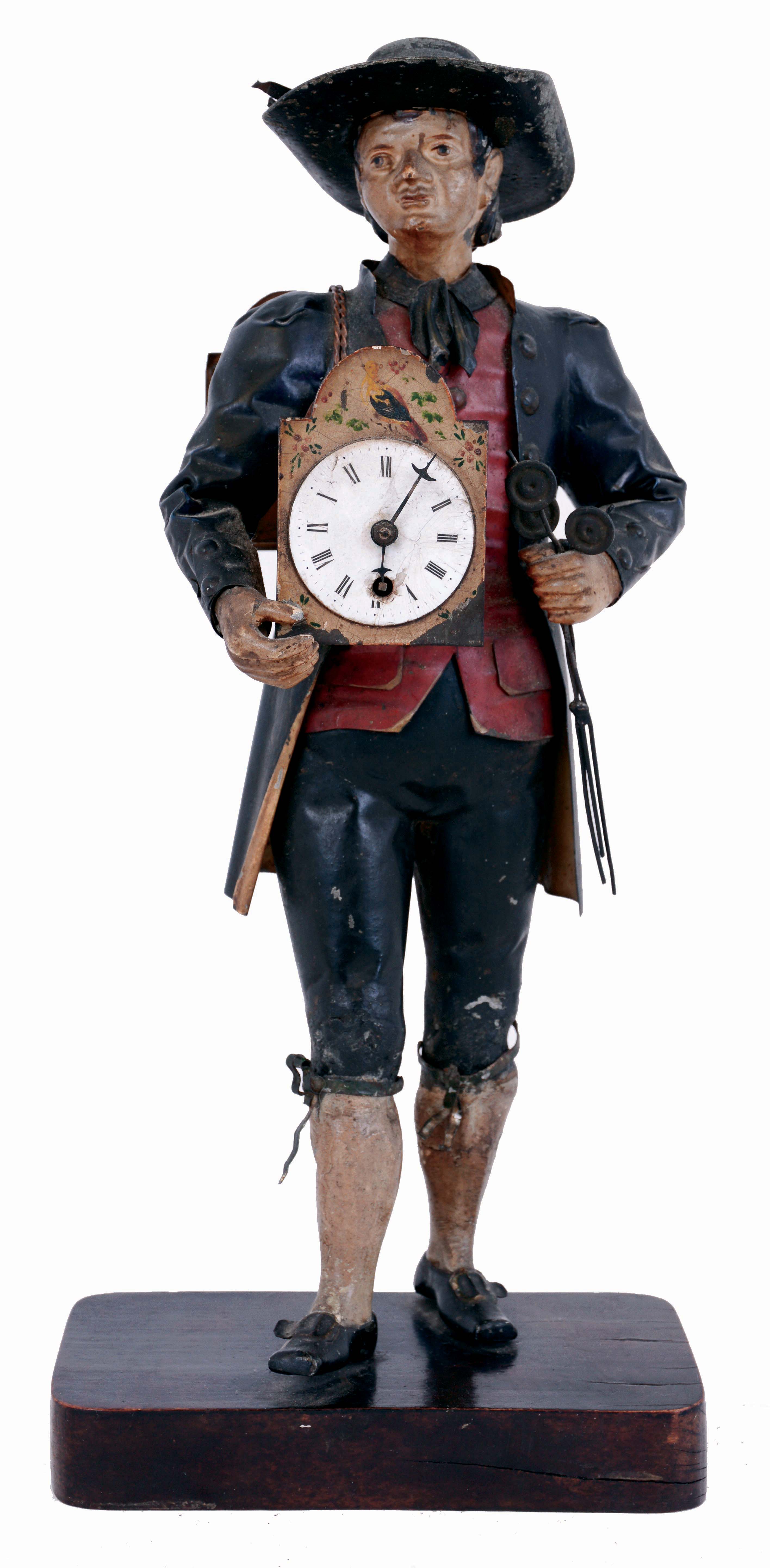 Germany, Black Forest "Uhrentrager" or clock peddler, the figure of polychrome painted sheet iron and in traditional dress, carrying a shield clock at his right, the clock with 30 hour pendulum timepiece, the back plate stamped "Schmutzer", Roman numeral white enamel dial and steel hands, also with another clock slung over his back with sliding front revealing a compartment for storing the key, his left hand clutching three pendulums, all mounted to wooden base, mid 19th century — Antique Clocks Price Guide