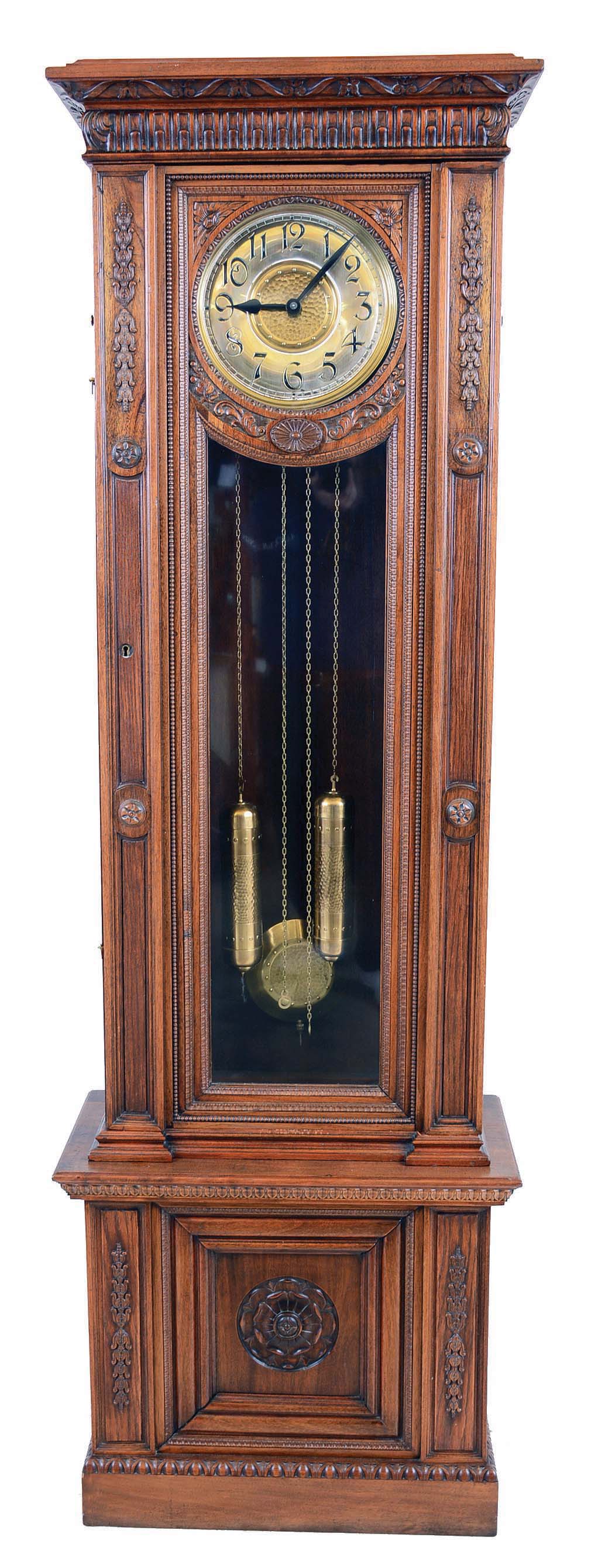 German, tall clock, 2-wt, 8-day time and strike, in a molded case with carved accents.  The round brass dial, embossed and with 2 added holes and arbor extensions for possible winding through the dial.  C.1915 — Antique Clocks Price Guide