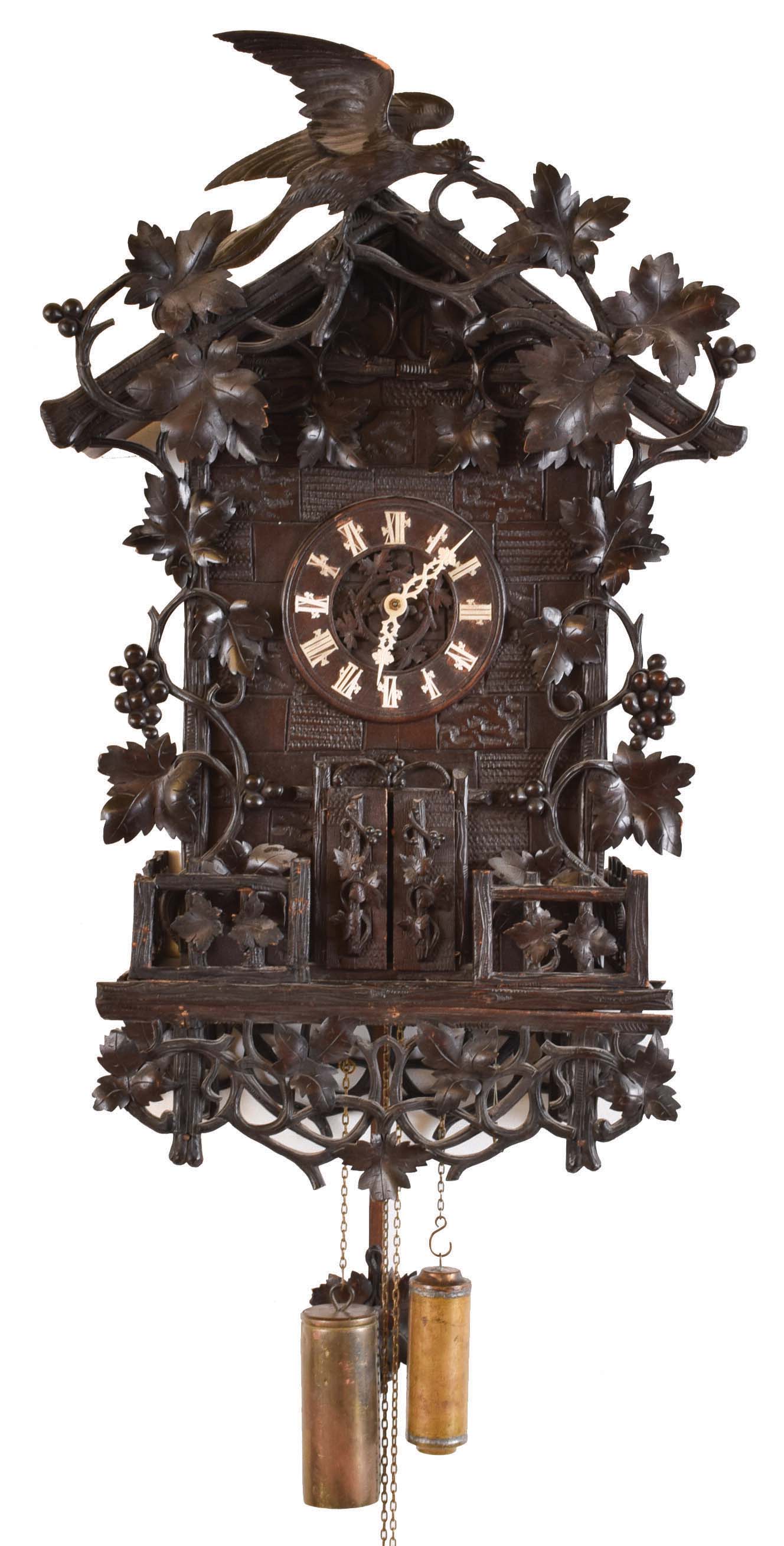 Black Forest hanging Flute Clock, the large carved case with carved vines, leaves and grapes, the top with similar ornament, and also a bird, the base with rustic railing, and with more trailing vines below, wooden dial with applied Roman numerals and carved bone hands, above the doors concealing the musicians, 30 hour, two train wood plate movement with brass wheels, the bellows mounted above, and with windchest and pipes at lower right, carved grape and leaf pendulum, brass weights — Antique Clocks Price Guide