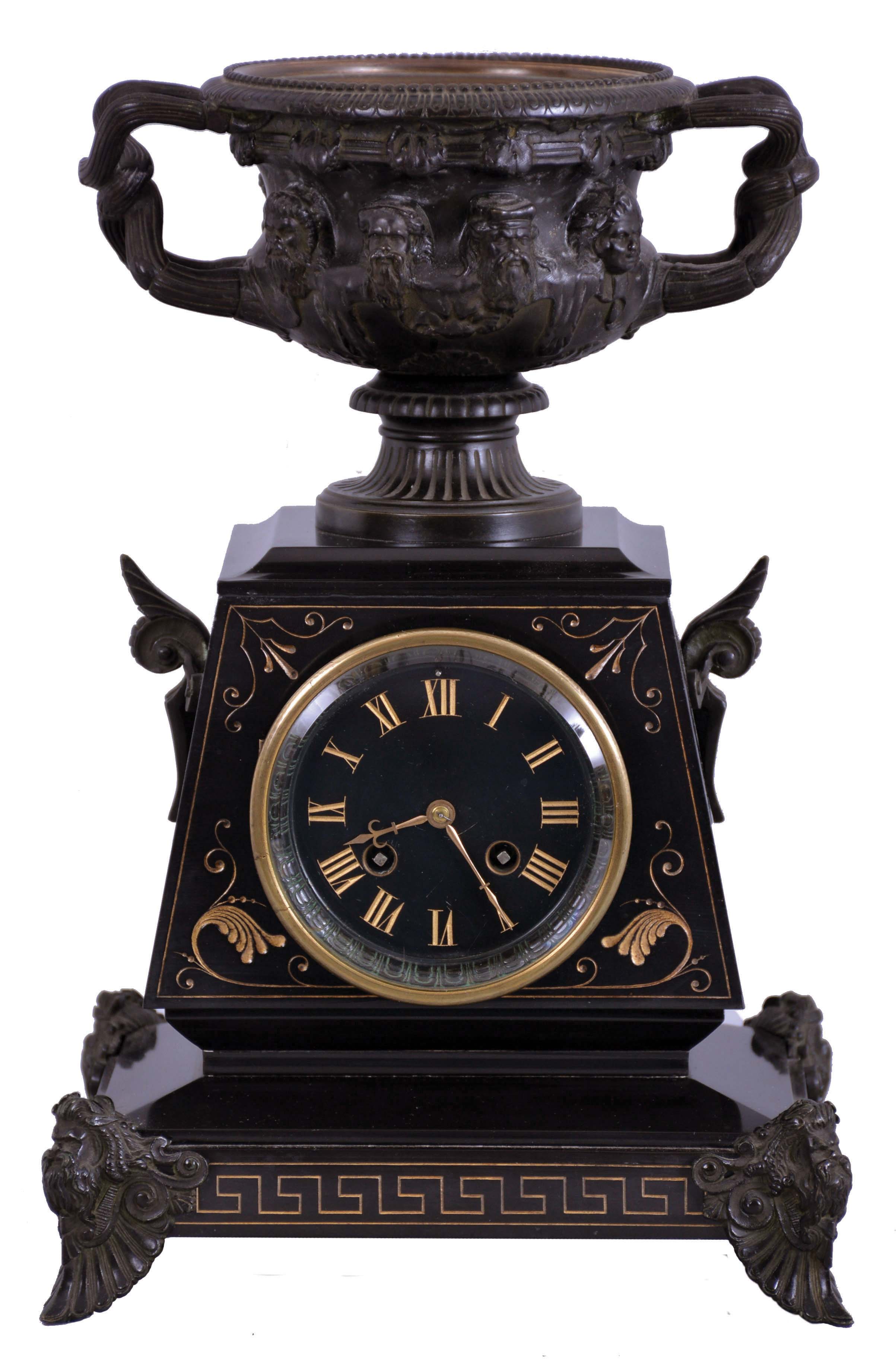 Charles Pickard, Paris, figural mantel clock, 8 day, time and strike movement by Japy Freres, patinated cast brass feet with human faces supporting a black slate case and dial with incised designs and numerals, surmounted by a handled urn displaying 8 human faces around the outside, c1880. — Antique Clocks Price Guide
