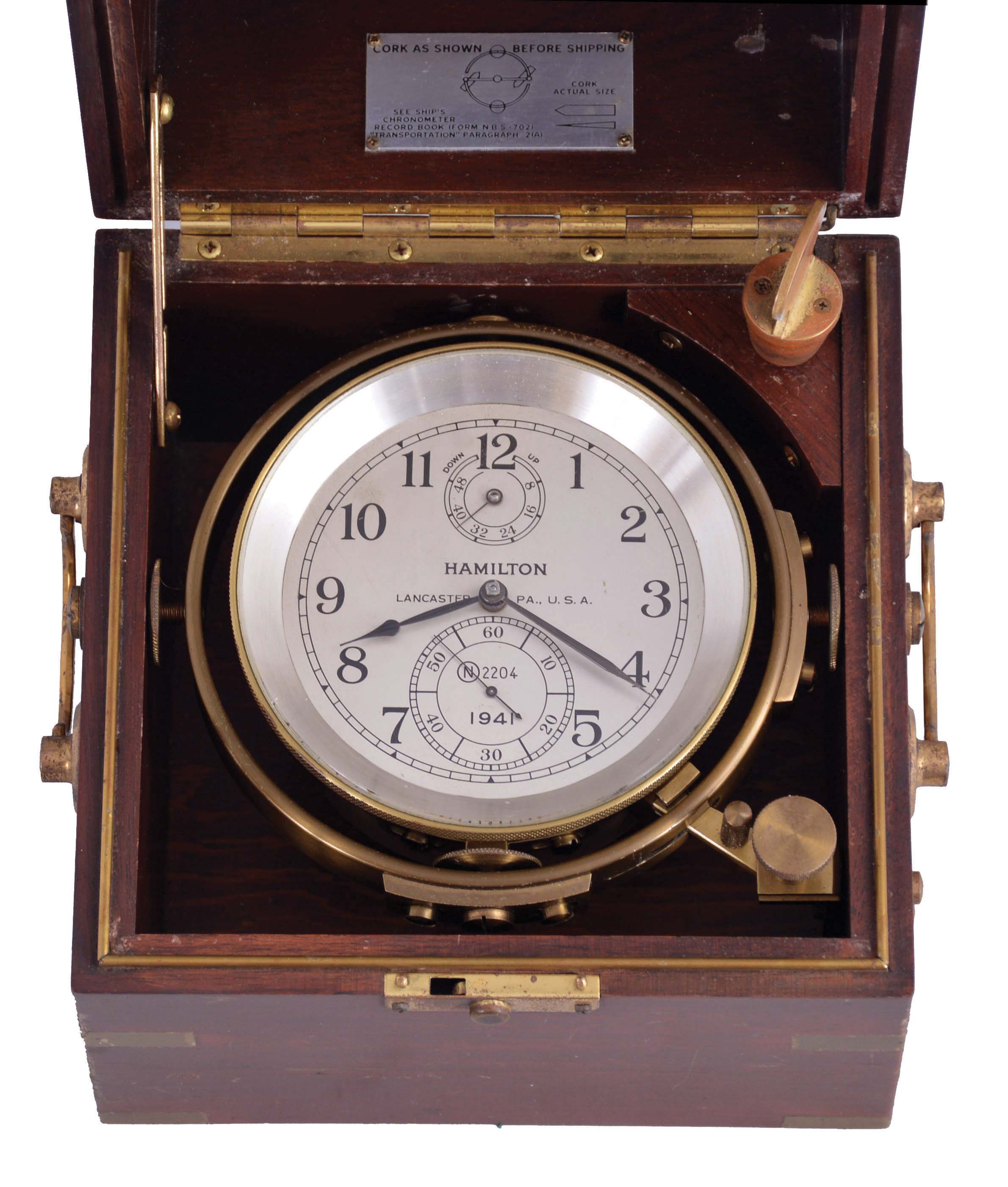 Hamilton Watch Co., Lancaster, Penn., model 21 marine chronometer, 14 jewel key wind and set, straight line damascened nickel plate movement with spring detent escapement and chronometer balance with helical hairspring, Arabic numeral metal dial with wind indicator, black painted steel spade and poker hands, contained in a gimbaled mahogany box with brass hardware, serial #2E2204, "N2204" marked on dial indicating that it was commissioned for use on a Navy vessel — Antique Clocks Price Guide