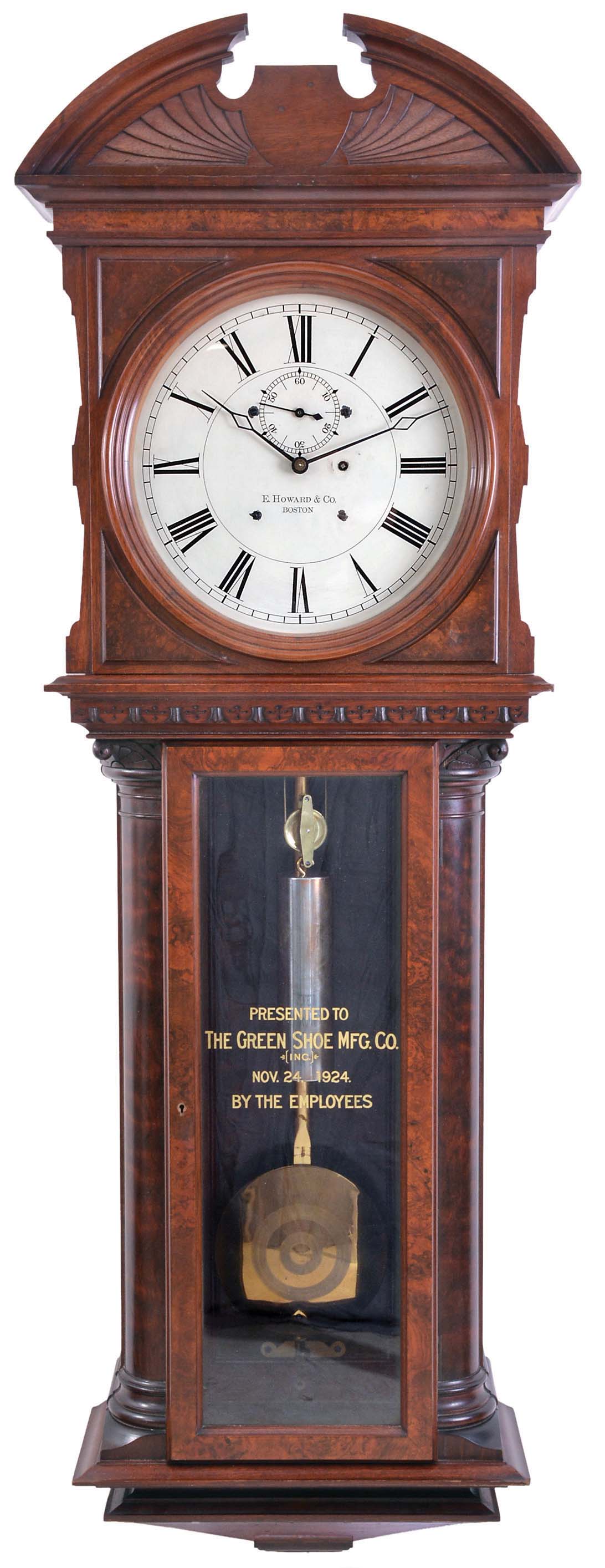 E. Howard & Co., Boston, Mass., "Regulator No. 57", jeweler's regulator wall clock, the walnut and burl walnut case with bold quarter columns, having turned and carved capitals and turned bases, flanking the burl veneer trunk door with reverse painted and gilt presentation, "THE GREEN SHOE MFG. CO. (INC.) NOV. 24, 1924. BY THE EMPLOYEES", over a burl veneered, stepped, tapered bottom bracket, upper door with raised wooden spandrels enframing the dial, over a nicely detailed carved molding above trunk door, and fan carved, broken arch top, Roman numeral painted dial signed "E. HOWARD & CO. BOSTON", 8 day, time only, weight driven movement with maintaining power and large brass, decorated bob and gold leafed pendulum rod, with cylindrical weight. — Antique Clocks Price Guide