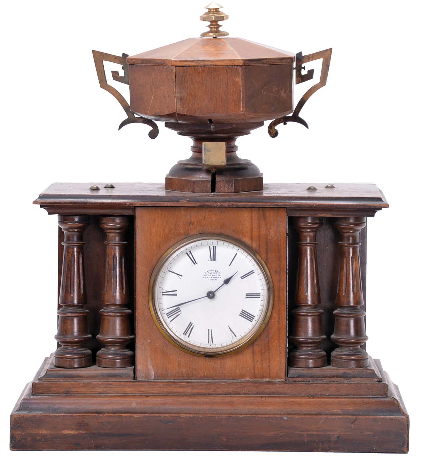 Automatic Memorandum Clock Co., London, "Davidson's Patent Automatic Memorandum Clock", 8 day time and reminder, spring driven, balance wheel movement in a walnut case with four full pillars and wooden urn on top. A time-only movement powers the clock and rotates a disk hidden within the urn. The disk has slots that correspond to different times of day. Memoranda, in the form of small bone tablets upon which one can write a few words, are placed within the slots. At the assigned time, a tablet falls through the slot, tips a brass tray, and triggers the battery powered alarm bell located in the base. The alarm would continue to sound until the tablet was removed. — Antique Clocks Price Guide