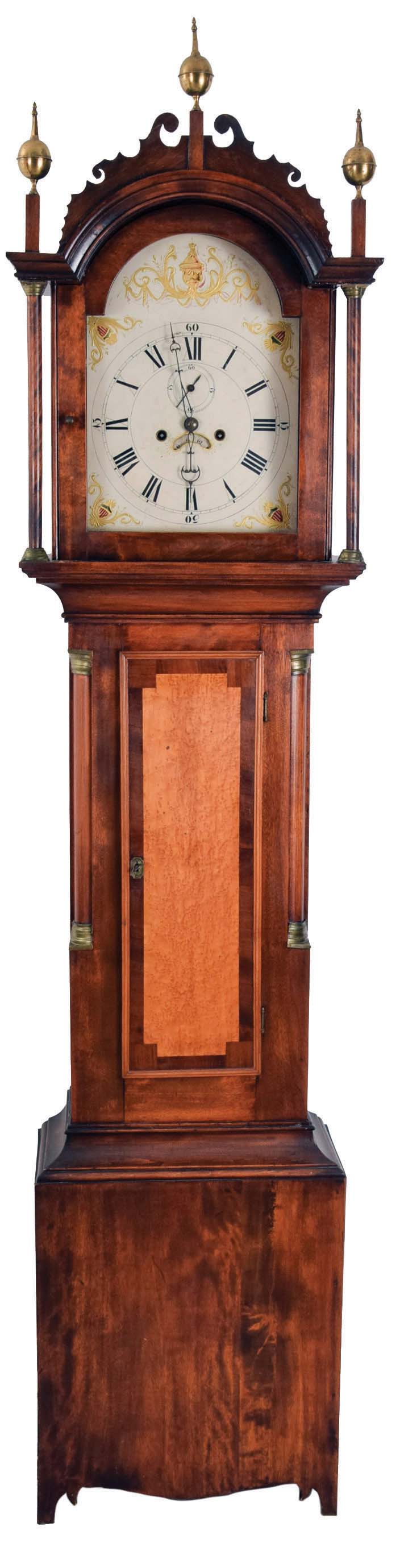 Tall case clock, New England, probably New Hampshire, flame birch case, trunk door with bird's eye maple panel framed with mahogany veneer. Quarter columns and brass capitals on the trunk, fret work crest, three brass finials and two full columns flanking hood. The associated movement is 8 day, time and bell strike, weight driven with brass plates and painted metal dial with calendar disc. — Antique Clocks Price Guide