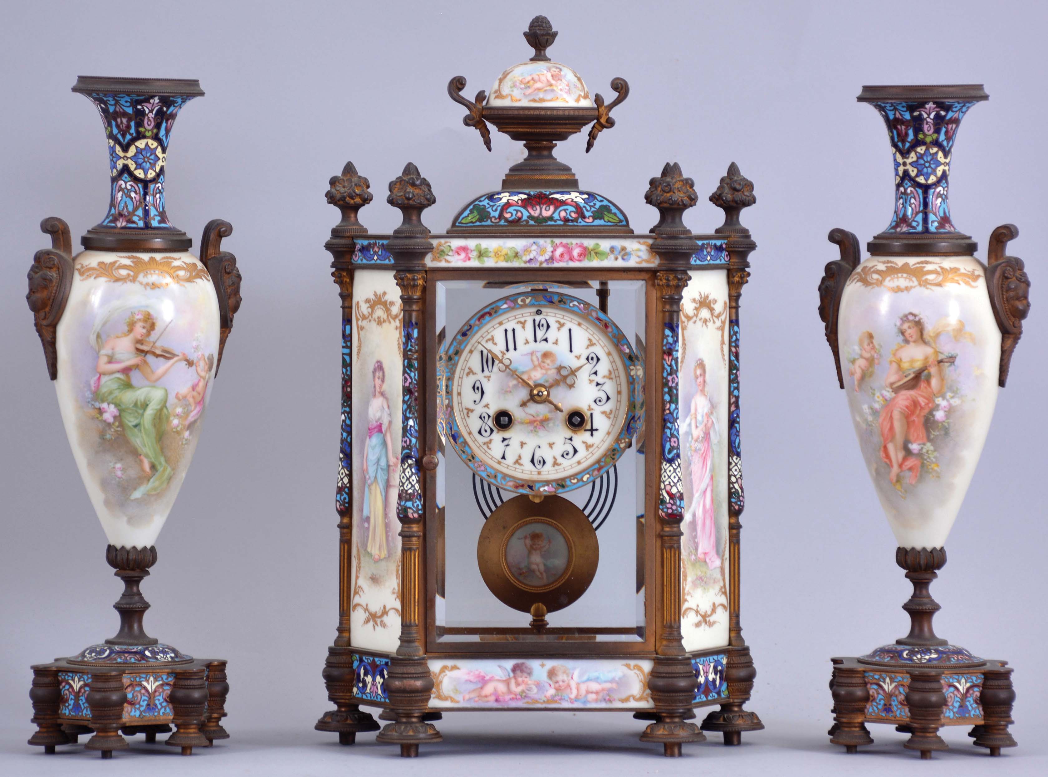 France, a decorative, three piece mantel garniture, the hexagonal, gilt bronze and champleve enamel clock case with columned corners, set with beveled glasses, and porcelain panels with polychrome enamel decoration, the panels with full length portraits of young women, floral ornament, and amorini, Arabic numeral white enamel dial with enamel painting of cupid, the pendulum also with his image, 8 day time and strike pendule de Paris movement, the tall, vasiform sidepieces with portraits and musical trophies, also with champleve enamel, matching the clock — Antique Clocks Price Guide