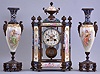 France, a decorative, three piece mantel garniture, the hexagonal, gilt bronze and champleve enamel clock case with columned corners, set with beveled glasses, and porcelain panels with polychrome enamel decoration, the panels with full length portraits of young women, floral ornament, and amorini, Arabic numeral white enamel dial with enamel painting of cupid, the pendulum also with his image, 8 day time and strike pendule de Paris movement, the tall, vasiform sidepieces with portraits and musical trophies, also with champleve enamel, matching the clock