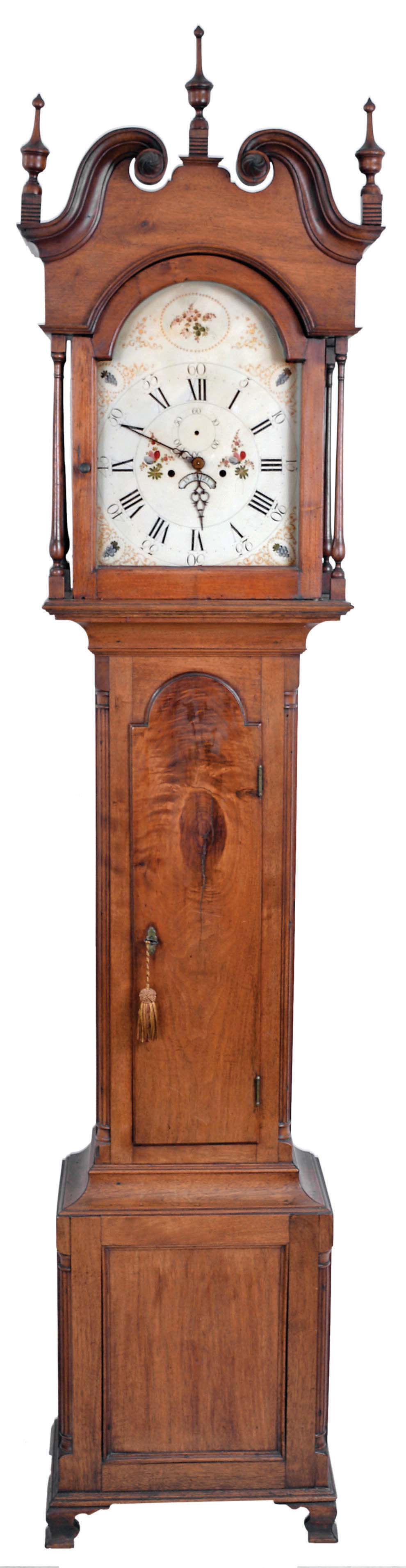 Pennsylvania tall clock, 30 hour, time and strike, weight driven brass movement in a walnut case with broken arch top, flame rosettes protruding about 1.5" from end of neck, three wooden finials on wooden chimneys, four shaped full columns flanking hood, paneled plinth, trunk door flanked by quarter columns is made with unusual large knot on ogee bracket base with quarter columns. The painted metal dial has calendar disc and floral and grape painted decorations. — Antique Clocks Price Guide