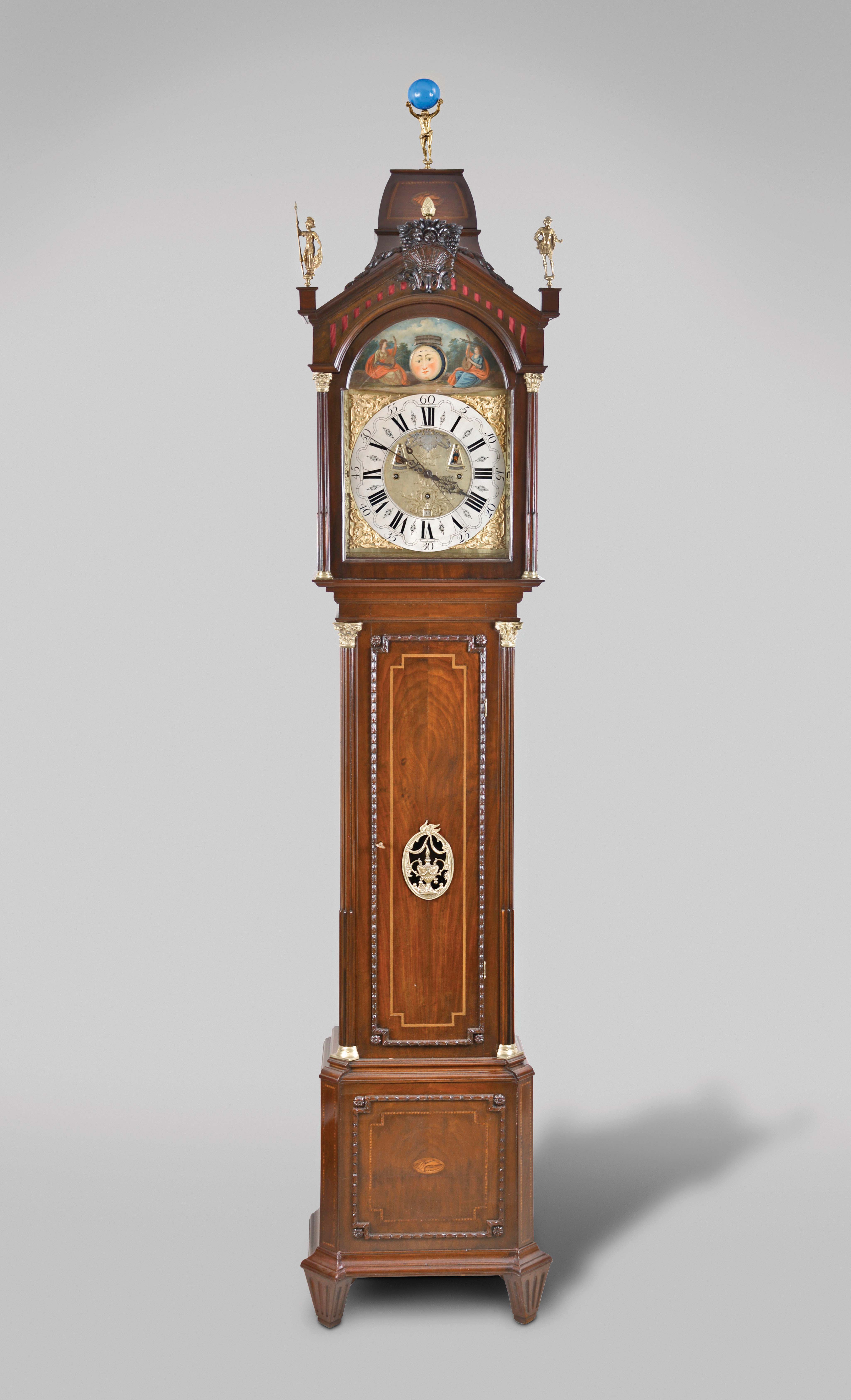 D.J. Tasma, Grouw, a monumental three train musical tall clock with full calendar, automata, and phases of the moon, mahogany case with molded base, fluted feet, canted corners, and carved bell flower molding with conforming marquetry band and central conch shell, the trunk with stop fluted half columns and Corinthian capitals, the door with carving and inlay matching the base, bonnet with stop fluted columns and Corinthian capitals below a pitched pediment with central carved basket of flowers, and three figural finials, brass composite dial with arcaded, silvered, roman numeral chapter ring, gilt spandrels, and engraved, matted center with calendar apertures, pierced blued steel hands, the arch with central moonphase aperture and polychrome painted landscape scene featuring two female musicians with lutes, their arms moving, as if to strum their instruments as the clock runs, and 8 day, three train, quarter striking musical movement, 7.5 inch cylinder playing 10 tunes on 12 bells, the quarters sounded on single bell, and striking the hours on a gong. — Antique Clocks Price Guide