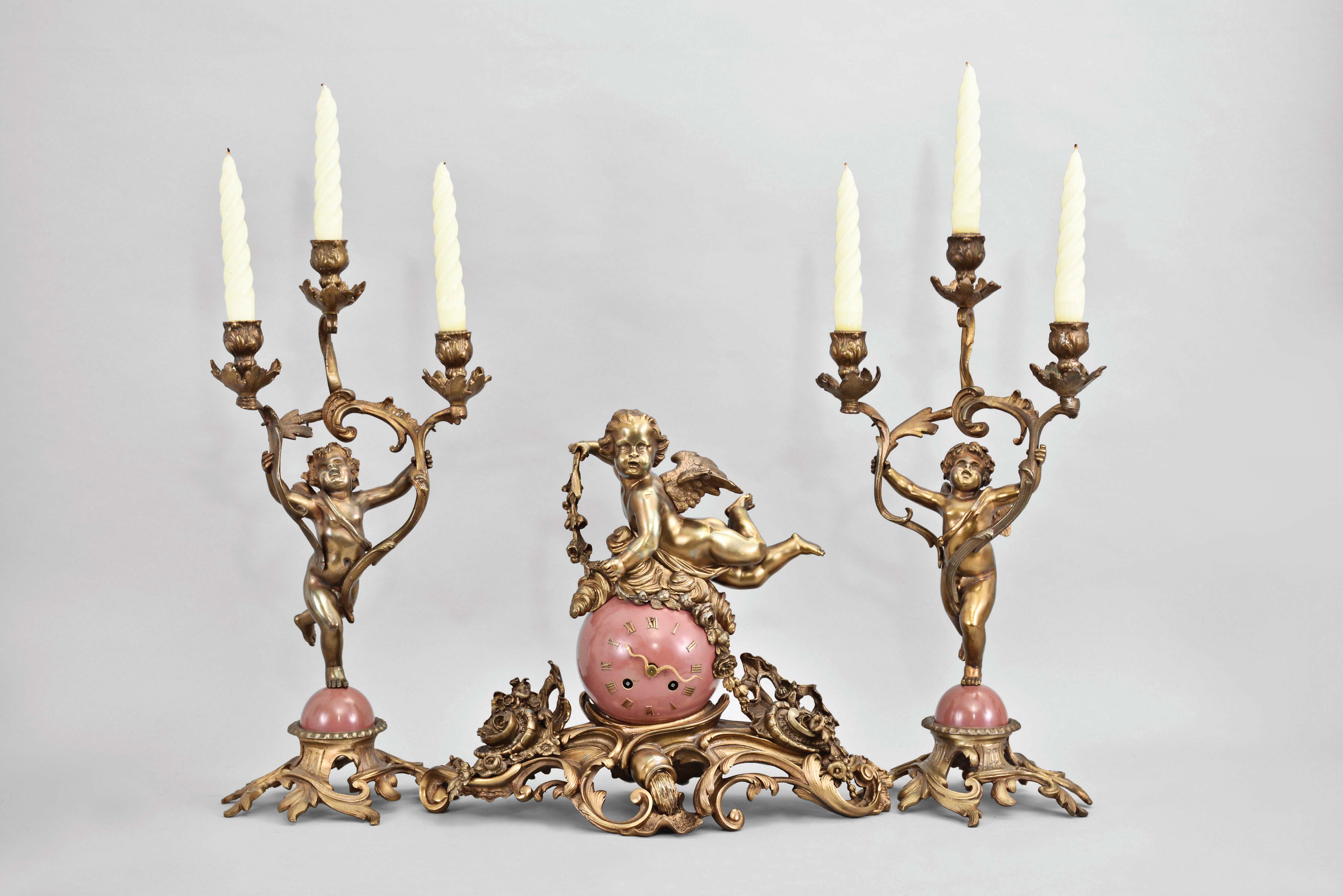 France, three piece mantel garniture set, 8 day, time and strike, spring driven pendule de Paris movement, the case with cast Rococo style base supporting a sphere with applied roman numerals, the hands in the form of a snake, all surmounted by a cast metal putto with outstretched wings holding a garland, the two matching side pieces with putti holding aloft the candleabra. — Antique Clocks Price Guide