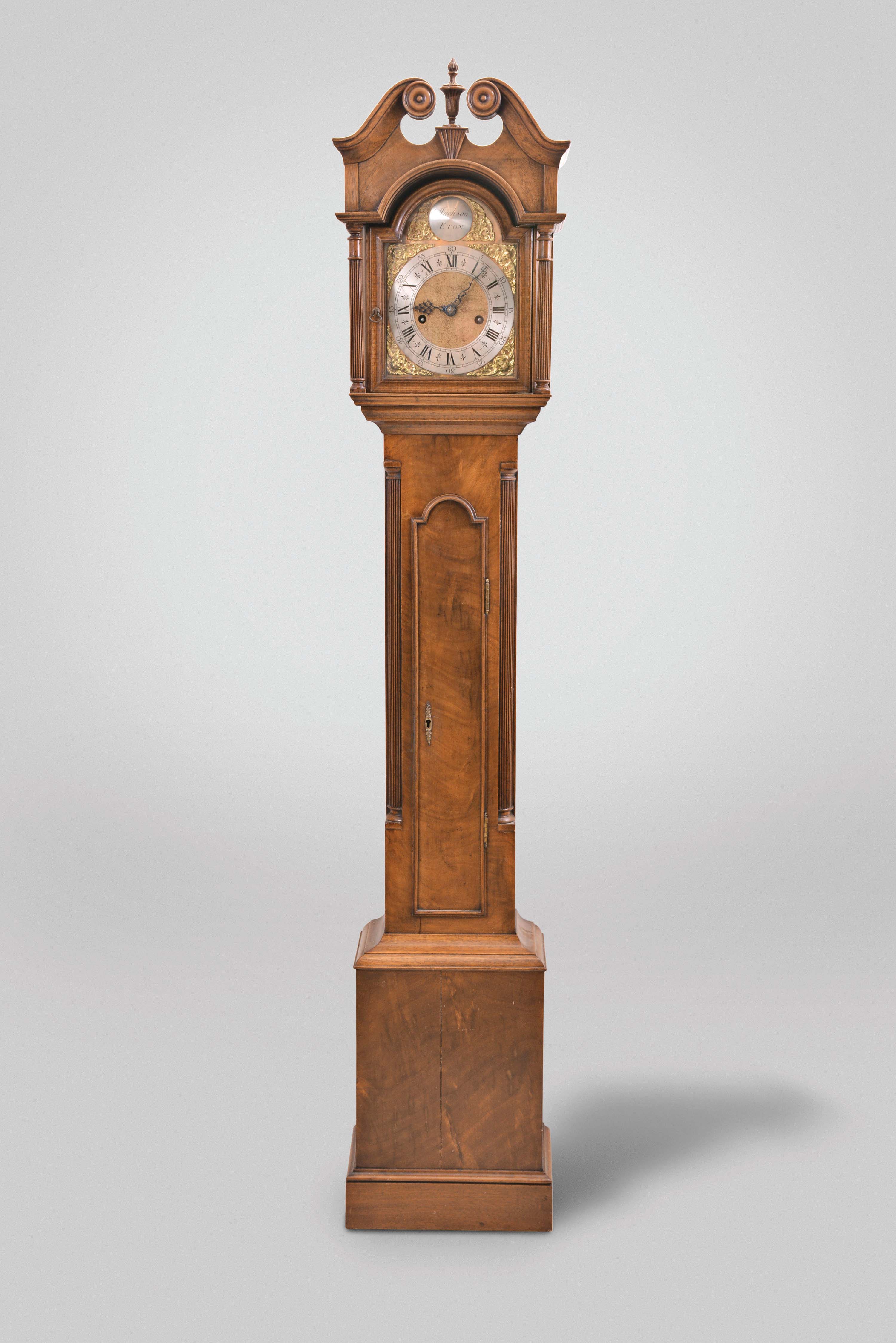 Reproduction dwarf tall clock, signed "JACKSON ETON", 8 day, time and strike, brass weight driven movement in a mahogany case with broken arch top, center flame finial above fluted plinth, tombstone door on trunk flanked by fluted columns and turned and fluted columns flanking hood door. The brass dial with silvered chapter ring has name boss and cast corner spandrels. — Antique Clocks Price Guide