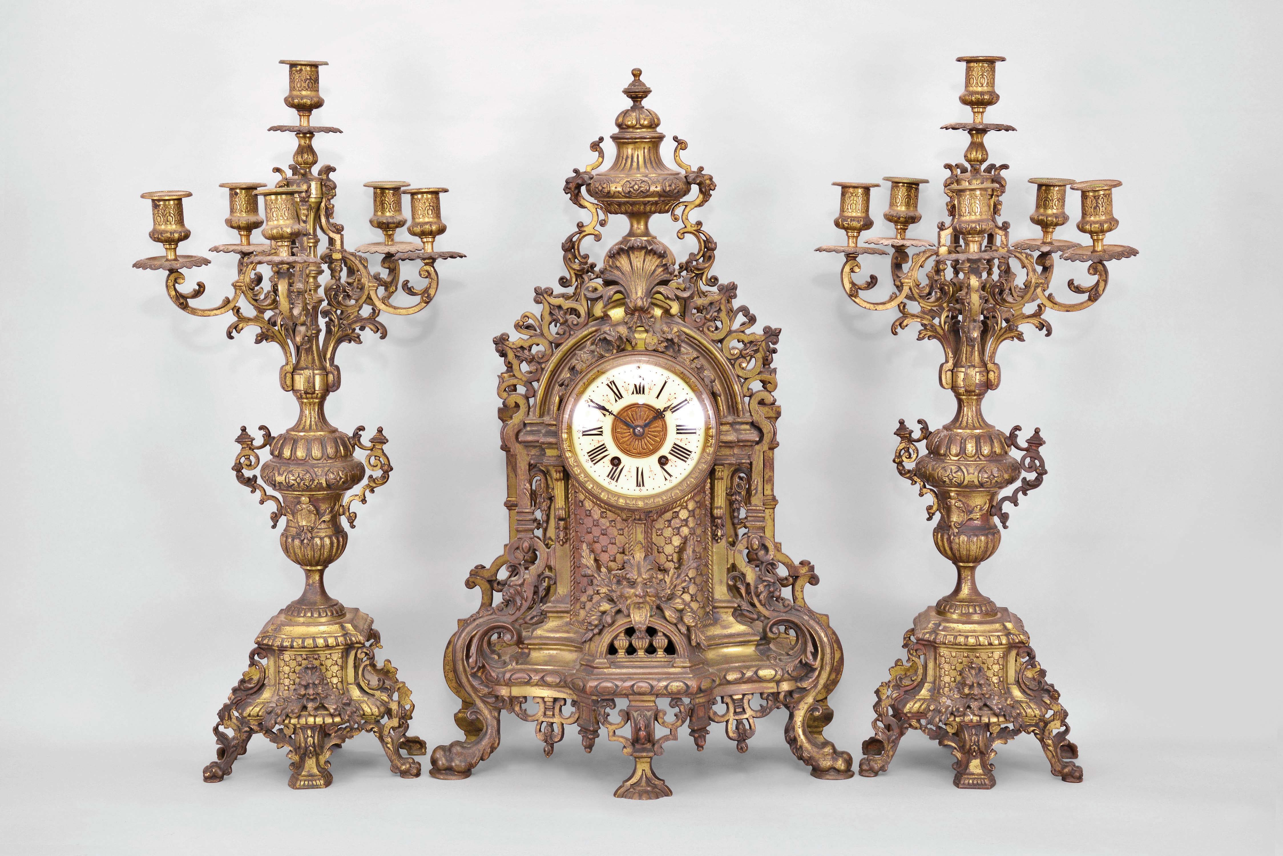 Jules-Edouard Dehon, Switzerland for Tiffany & Co., New York, three piece mantel garniture set, 8 day, time and strike, spring driven movement in a elaborately sculpted cast brass Renaissance revival case featuring claw feet, foliate heads, floral motifs, peripheral fretwork, and lion masks in profile, cream porcelain dial with blued steel hands, all topped by a large urn finial. Two matching brass candelabra holding six candles each. — Antique Clocks Price Guide
