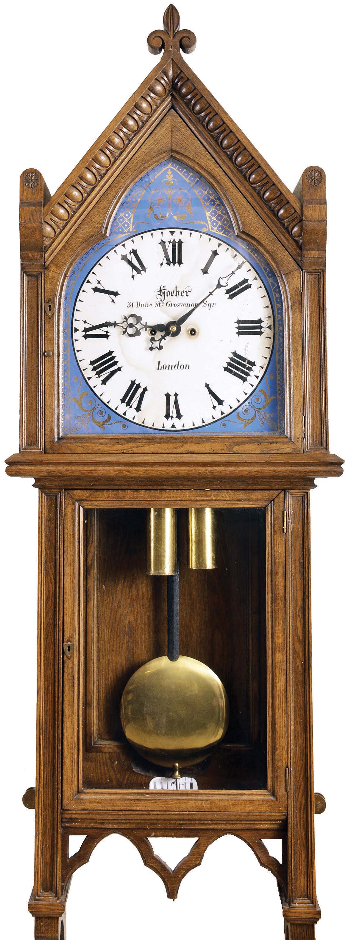 Louis Hoeber, 31 Duke St, Grosvenor Square, London, England, monumental Gothic revival wall clock, 8 day, time and strike, weight driven movement in an oak case with fleur-de-lis pediment top, and egg and dart molding flanked by pinwheel carved side elements over glazed door exposing large brass pendulum bob and beat scale. The reverse painted glass Gothic roman numeral dial with gilt lattice surround on a blue ground has maker's signature, address and Gothic hands — Antique Clocks Price Guide