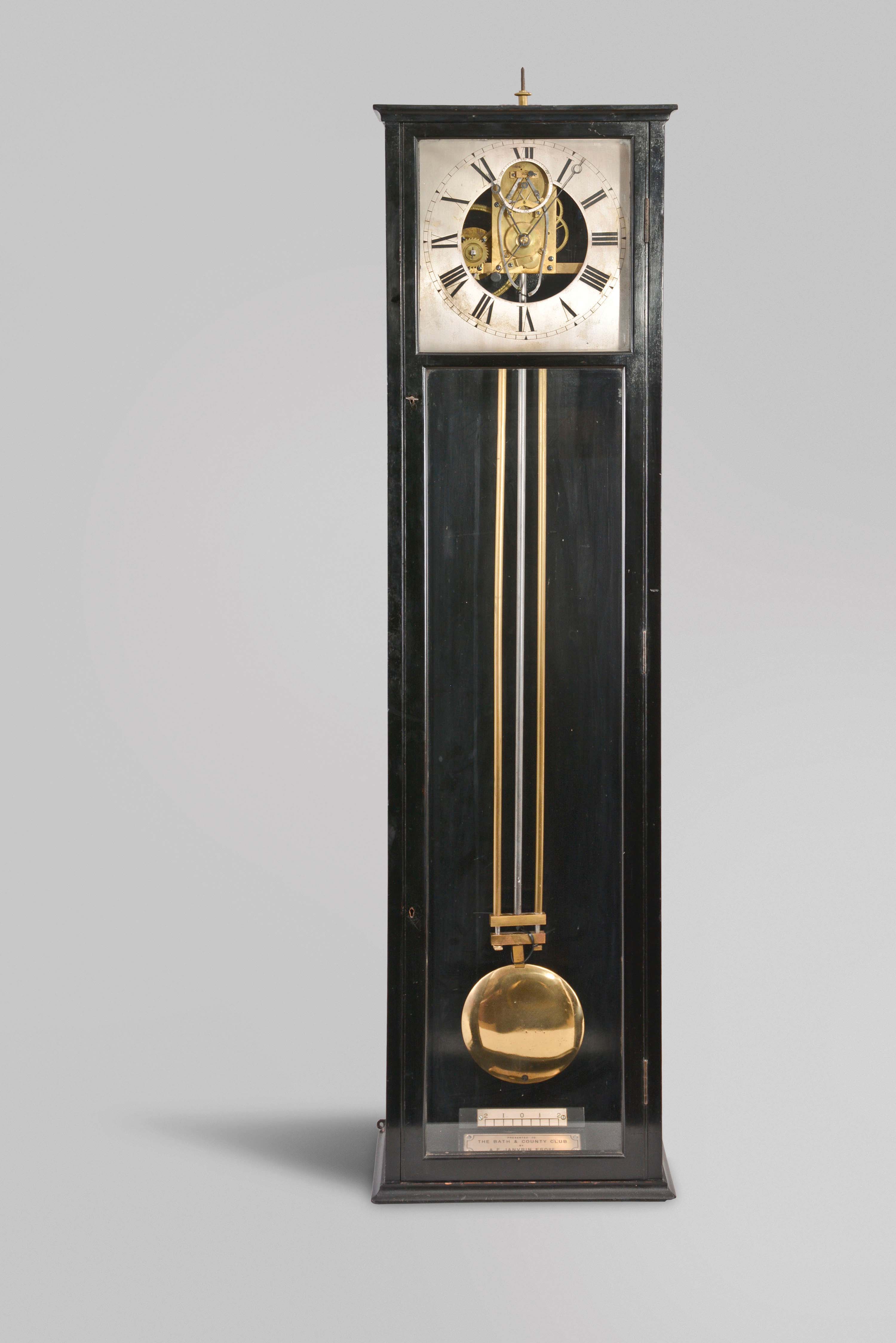 England, an ebonized hanging regulator with double three legged gravity escapement, the rectilinear four light case with simple molded top and base, the bottom with beat scale and presentation plaque "Presented to the Bath and Country Club A.F. Janvrin Esq.", roman numeral silvered dial with open center, blued steel Breguet style hands, brass, spring driven movement mounted to a heavy, cast iron bracket, three rod gridiron pendulum with brass bob — Antique Clocks Price Guide