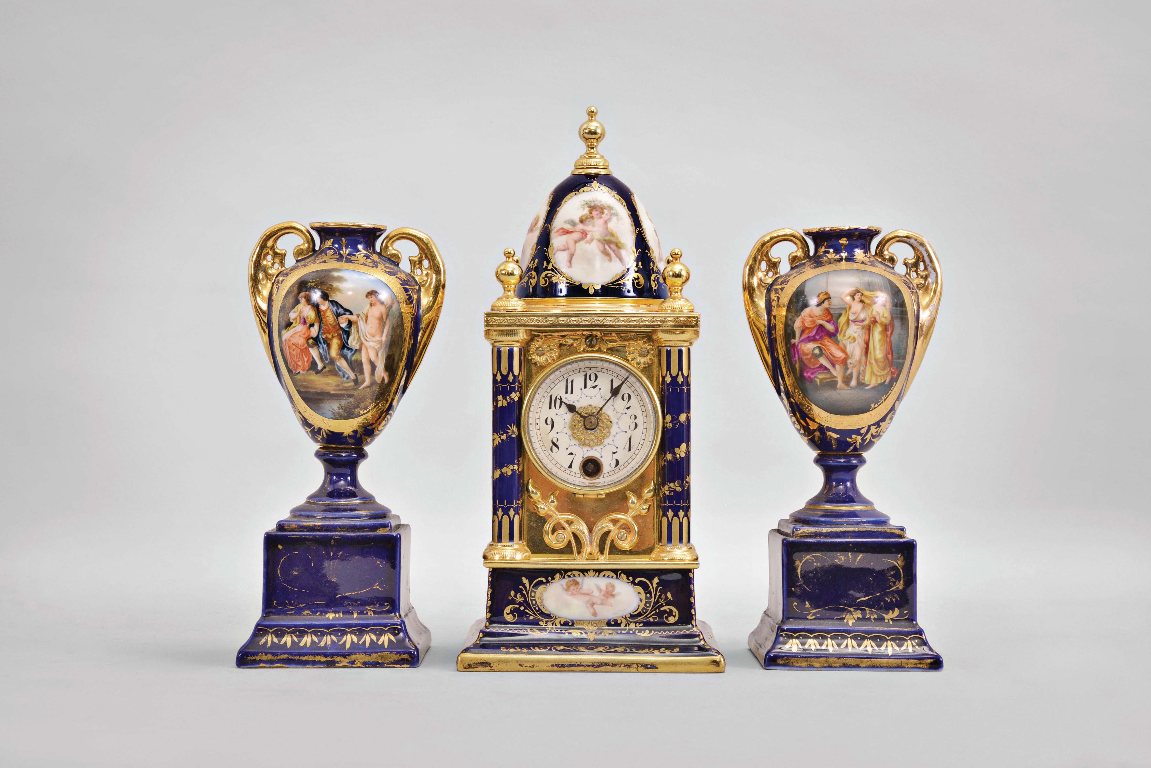 Austria, a Royal Vienna porcelain three piece garniture, the architectural clock case with cobalt blue ground, gilt, foliate ornament, and vignettes featuring putti in the clouds, gilt brass mid section surrounded by six porcelain columns, and containing the 8 day, timepiece movement, Arabic numeral white enamel dial, blued steel spade hands, and matching vasiform side pieces on square plinths, also in cobalt with gilt ornament, and each with courting scene in an oval reserve, signed "Hauffmann", all bases with beehive mark — Antique Clocks Price Guide