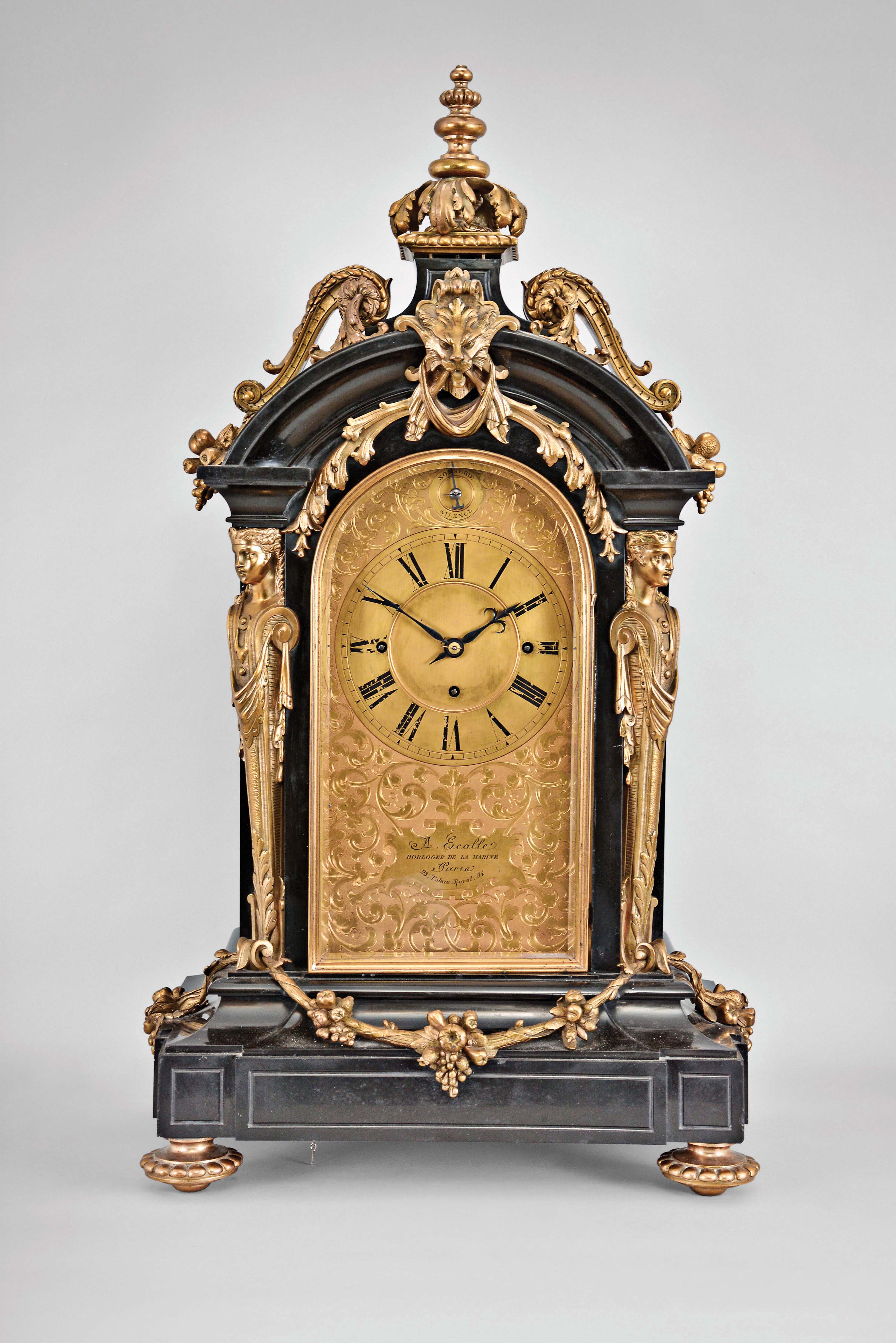 A. Ecalle, Horloger de la Marine, 93- 94 Palais Royal, Paris, a monumental Belgian slate, gilt bronze mounted, grand sonnerie mantel clock, on gilt, gadrooned feet, the base with incised panels and deep cavetto molding, and hung with a gilt, fruit and foliate festoon, the recessed corners with gilt caryatids supporting the cavetto molded cornice and arch top, the arch with central lion mask and foliate ornament issuing from its mouth, the top with gilt scrolls flanking a stone plinth surmounted by a turned, gadrooned and acanthus ornamented finial, engraved, gilt dial with scrolling foliage, Roman numeral chapter ring and subsidiary sonnerie/silence selector, and with retailers name and address within a blank reserve below the dial, finely cut and finished blued steel hands, and massive three train, quarter striking fusee movement, sounding the quarters and hours on five gongs — Antique Clocks Price Guide