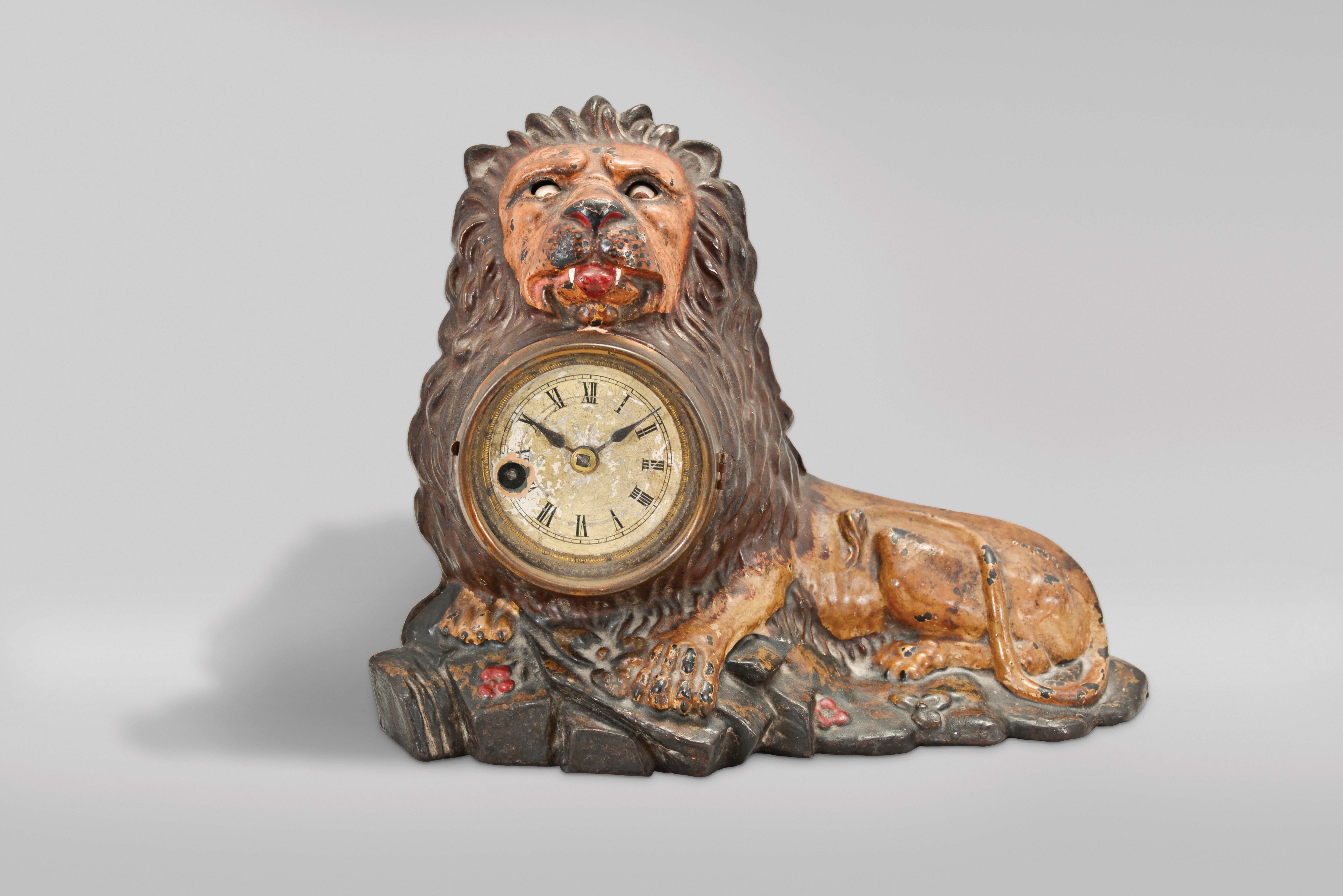 Bradley & Hubbard, Meriden, Conn., "Lion" blinking eye shelf clock, the polychrome painted, cast iron beast recumbent in a rocky landscape and appearing to hold the bow of a giant pocket watch in his mouth, his eyes connected to a lever on the pallet arbor, the rocking arbor imparting an up and down motion to the eyes, roman numeral, varnished paper dial with blued steel spade hands, and 30 hour pendulum timepiece. — Antique Clocks Price Guide