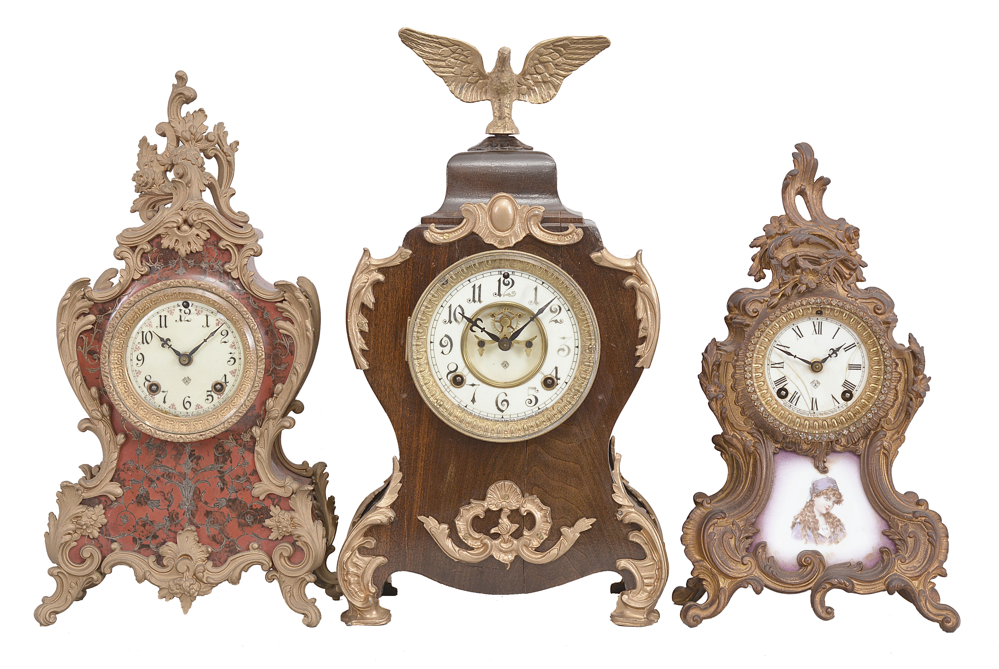 Clocks- 3 (Two) Ansonia Clock Co., New York, 8 day, time and strike, spring driven movements, porcelain dials: (1) "Apollo" mantel clock finished in red with gilt trimmings, c1901; (2) "Triano" mantel clock mantel clock with purple painted porcelain panel, c1901; (3) Mantel clock in a wood case with applied cast ornaments and eagle finial, c1890. — Antique Clocks Price Guide