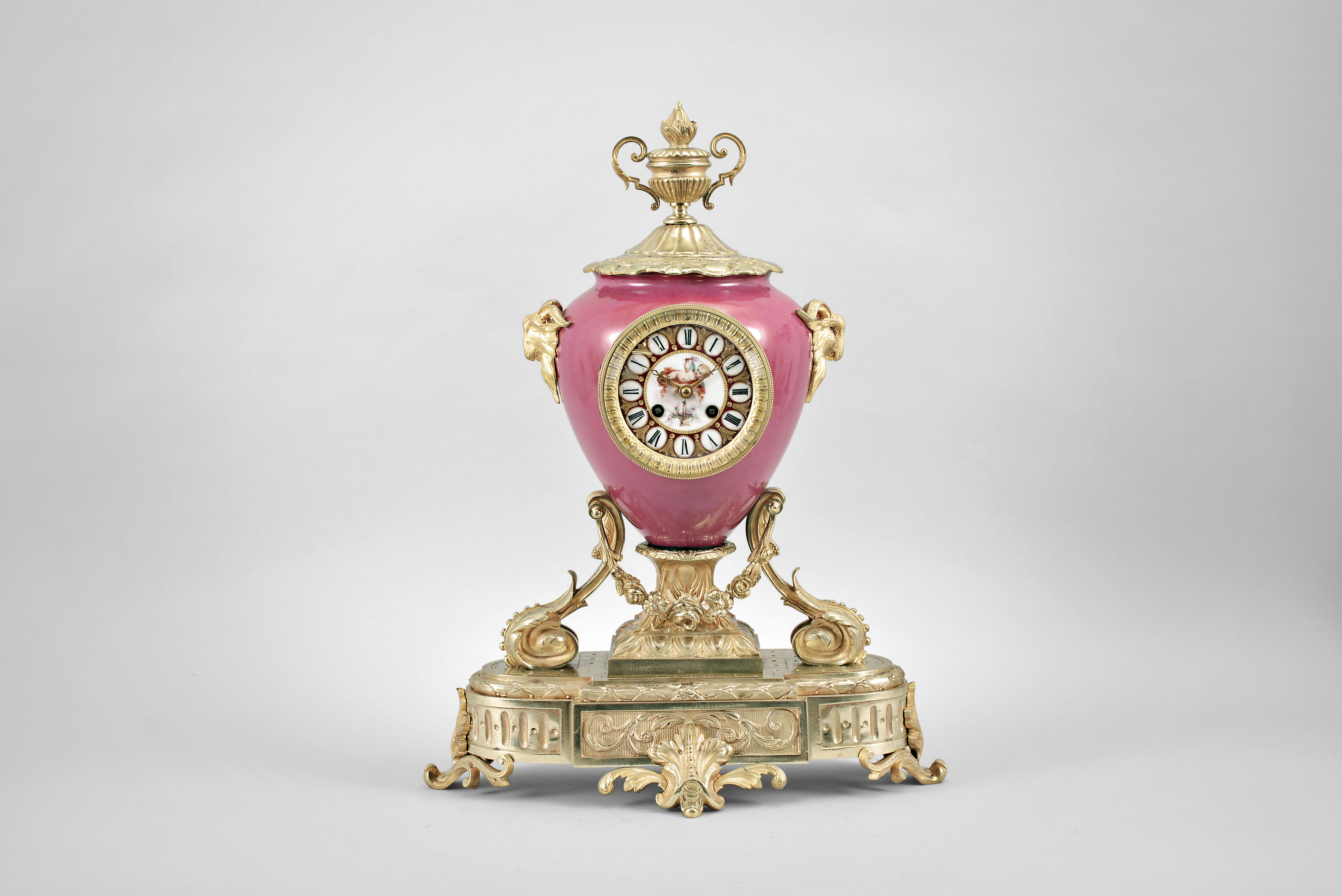 France, ceramic and ormolu mantel clock, 8 day, time and strike, spring driven pendule de Paris movement in a vibrant porcelain vase-form case mounted on a gilt metal base with floral swag, flanked by horned rams, and topped with an urn finial, polychrome painted dial with putto, gilt hands. — Antique Clocks Price Guide