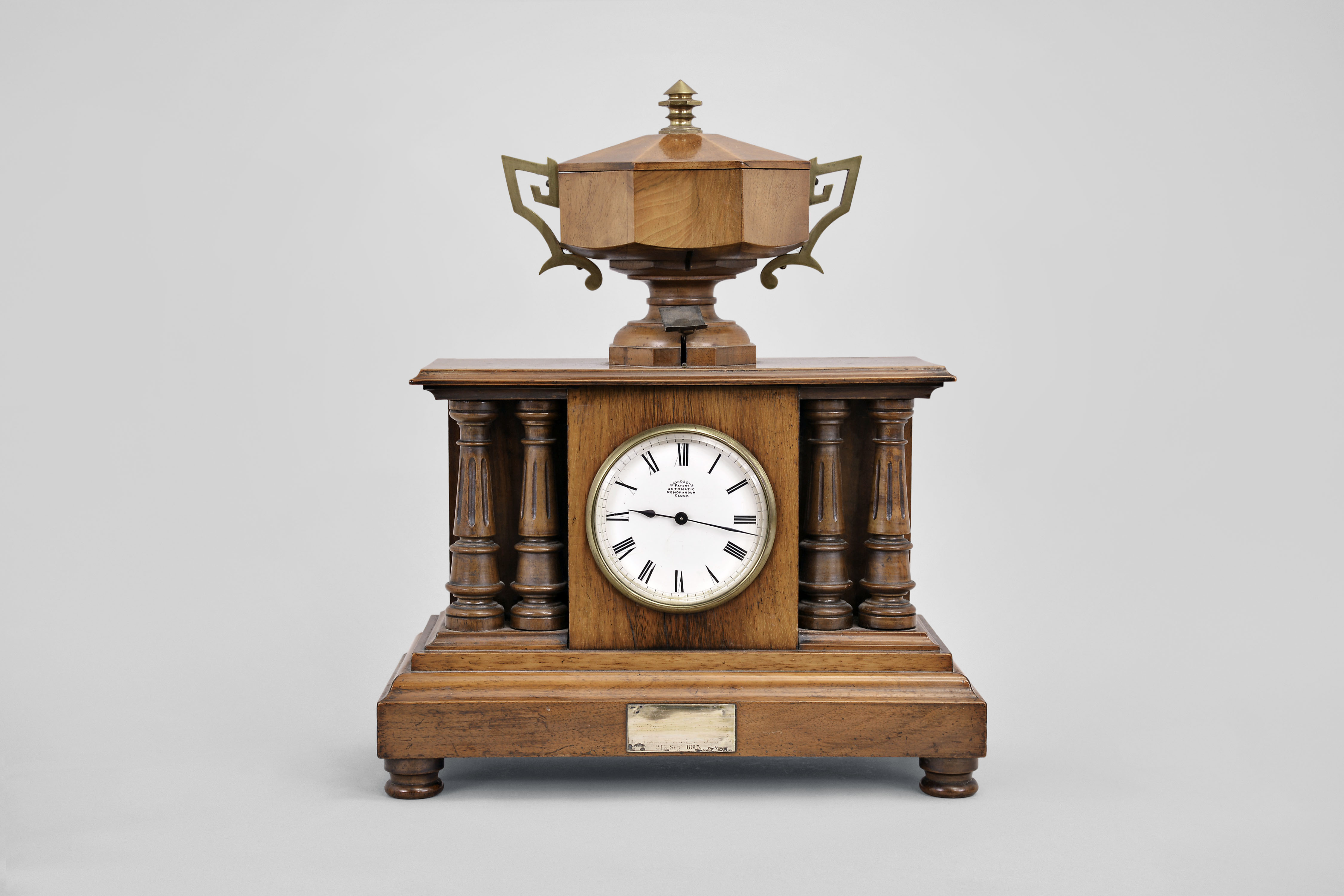 Automatic Memorandum Clock Co., London, "Davidson's Patent Automatic Memorandum Clock", 8 day time and reminder, spring driven balance wheel movement in a walnut case with four full pillars and wooden urn on top, white enamel dial and blued steel hands. — Antique Clocks Price Guide
