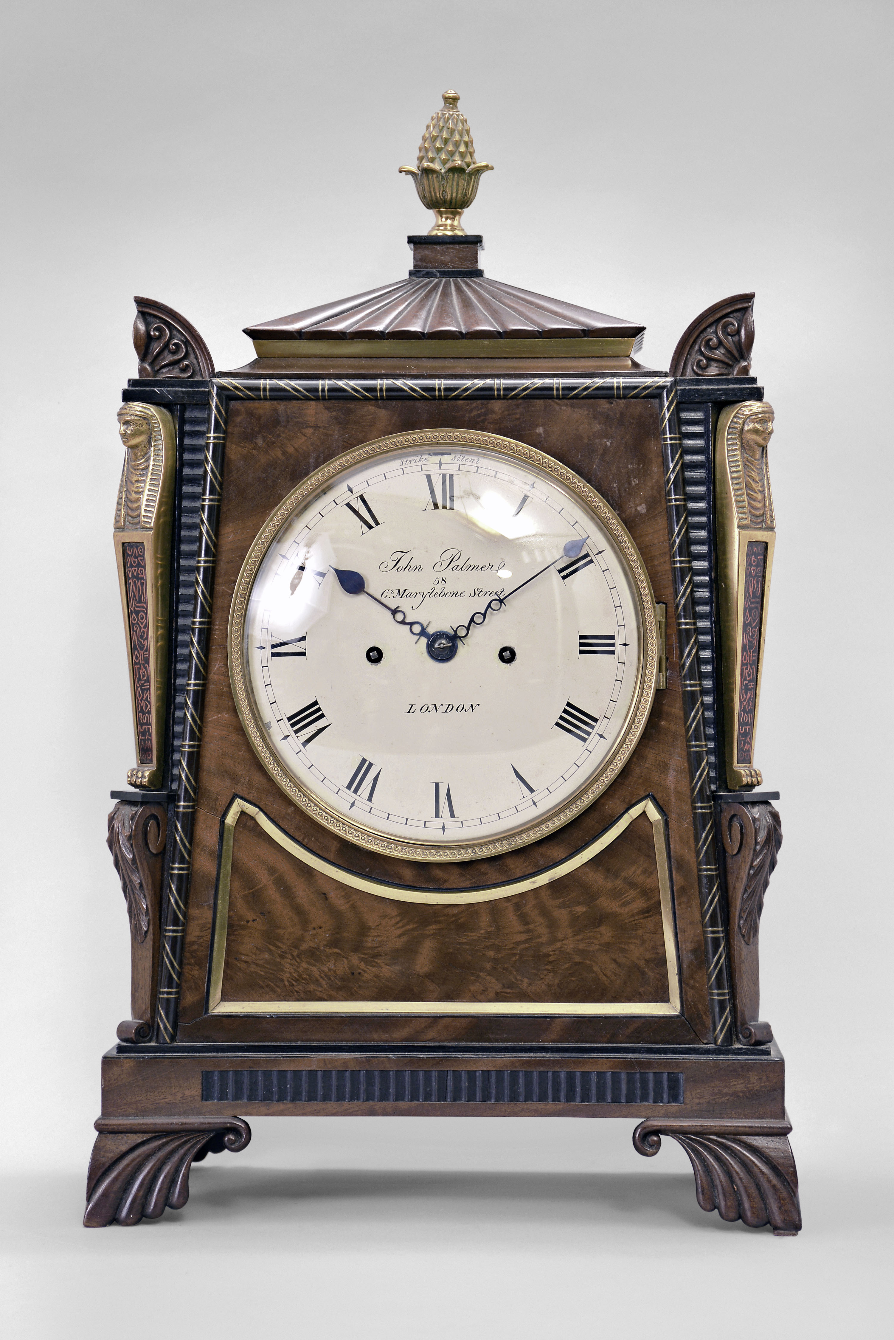 John Palmer, London, bracket clock, 8 day, time and strike, spring driven fusee movement in an Egyptian Revival case made of mahogany with brass inlay and applied brass figures on the corners, the top with anthemion corners, gadrooned plinth supporting a brass finial, painted metal dial signed as above, blued steel spade variant hands. — Antique Clocks Price Guide