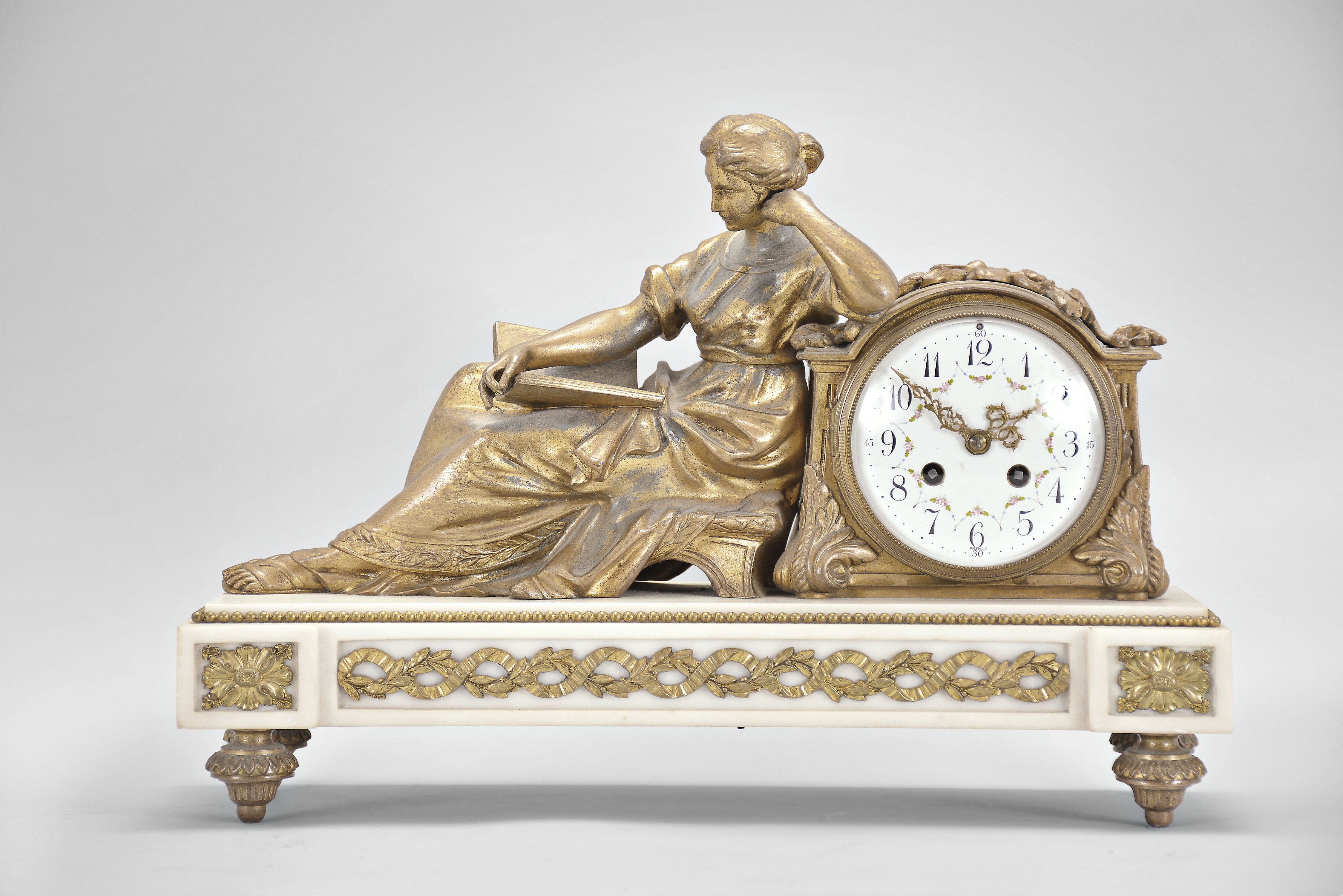 France, figural mantel clock, 8 day, time and strike, spring driven pendule de Paris movement in a cast metal case mounted on a white marble base with cast gilt feet and applied ornaments, the clock with white enamel dial, polychrome garlands, and Louis XV style hands, all adjacent to the figure of a seated woman reading a book. — Antique Clocks Price Guide